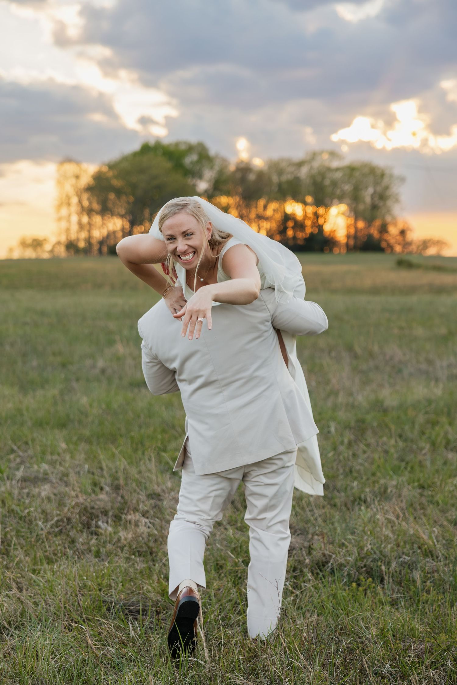 Groom carrying laughing bride on his back through an open field at golden hour sunset.
