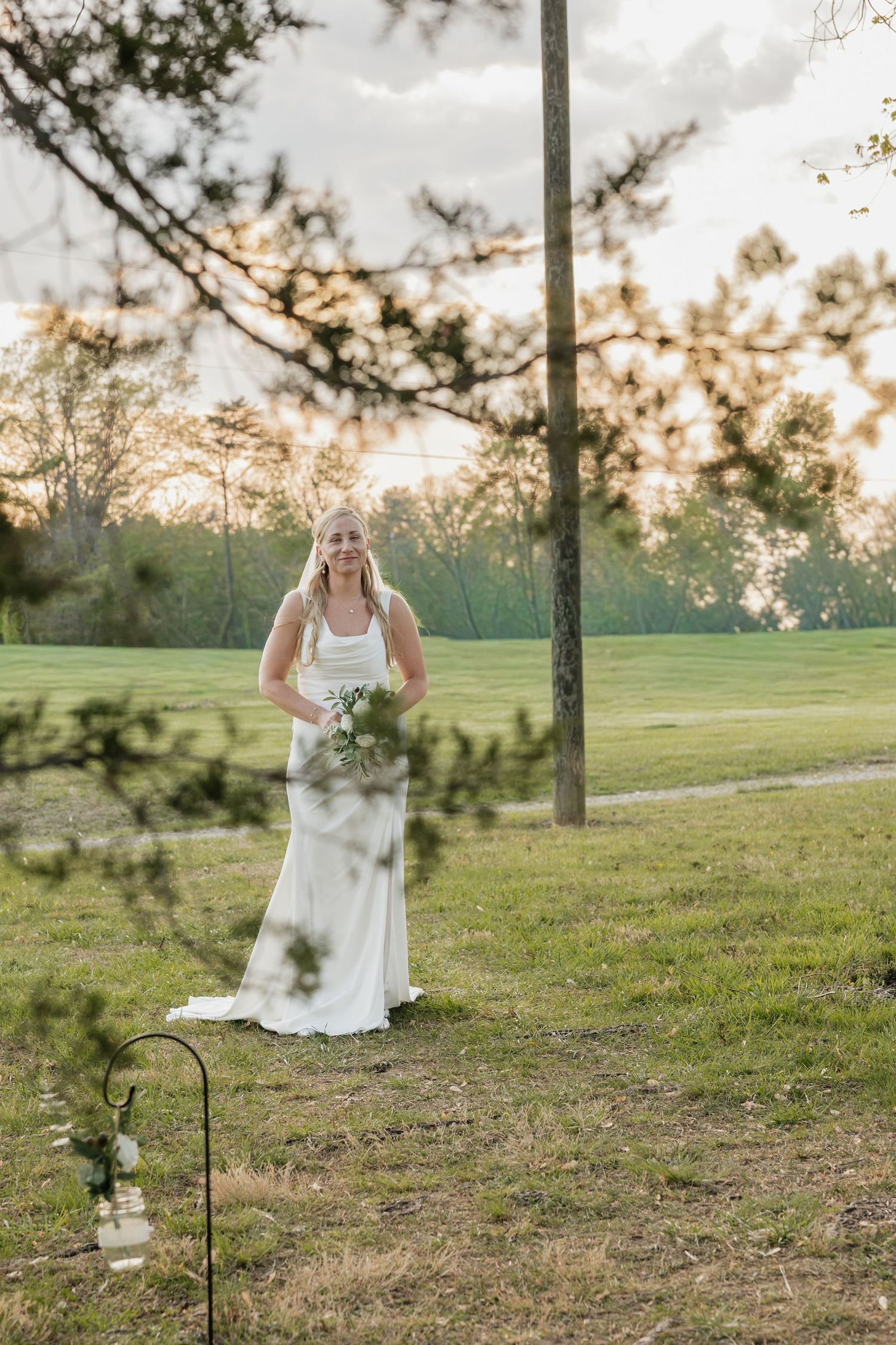 Beautiful bride in white gown holding bouquet walking toward outdoor wedding ceremony at golden hour.