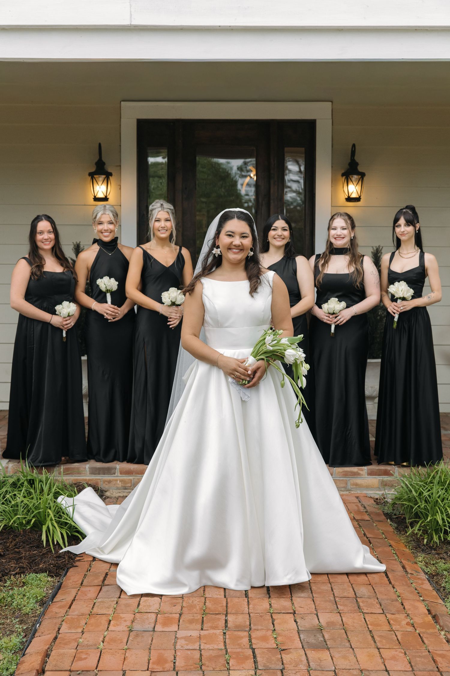 Smiling bride in white ball gown stands before bridesmaids in black dresses holding white floral bouquets.