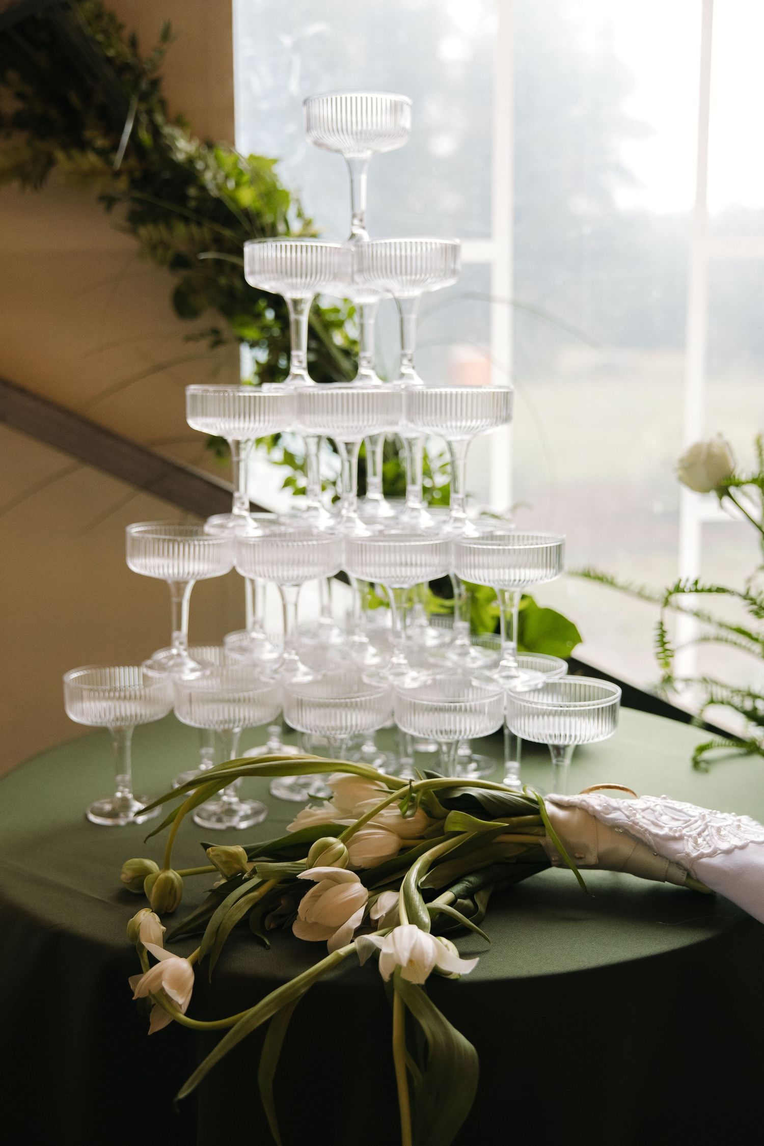 Crystal champagne tower on green tablecloth with white tulip bridal bouquet and greenery backdrop.