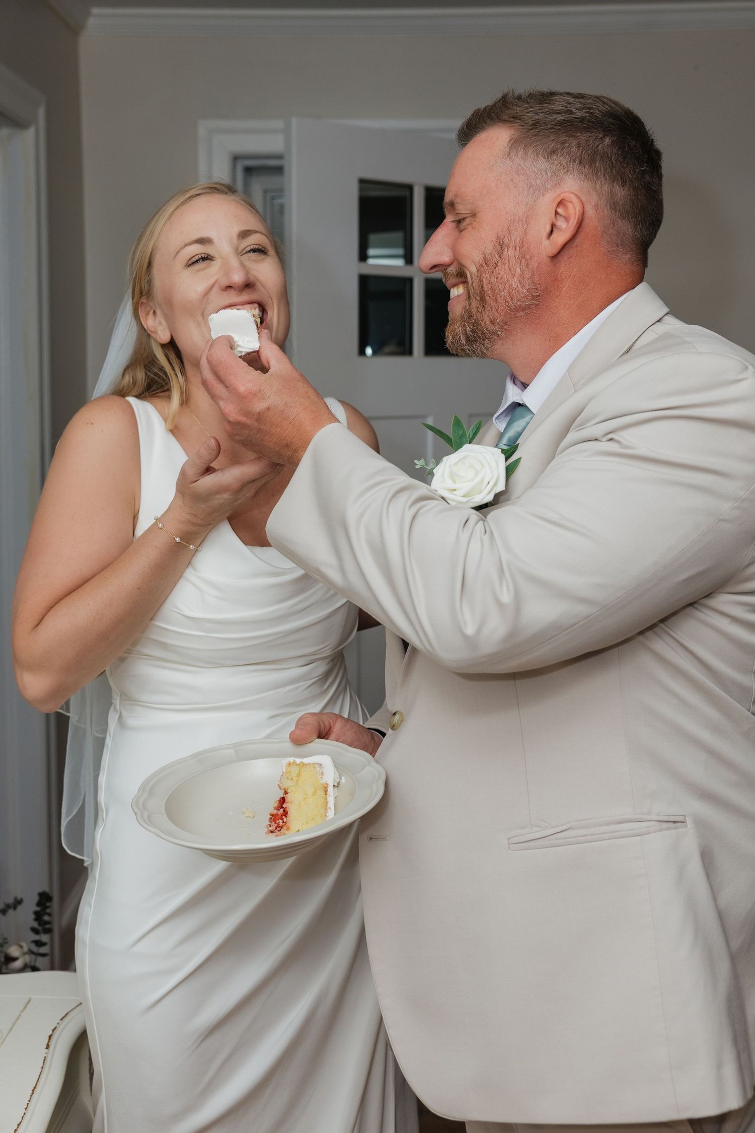 Groom feeds bride a bite of wedding cake while she laughs joyfully, holding a plate of cake.