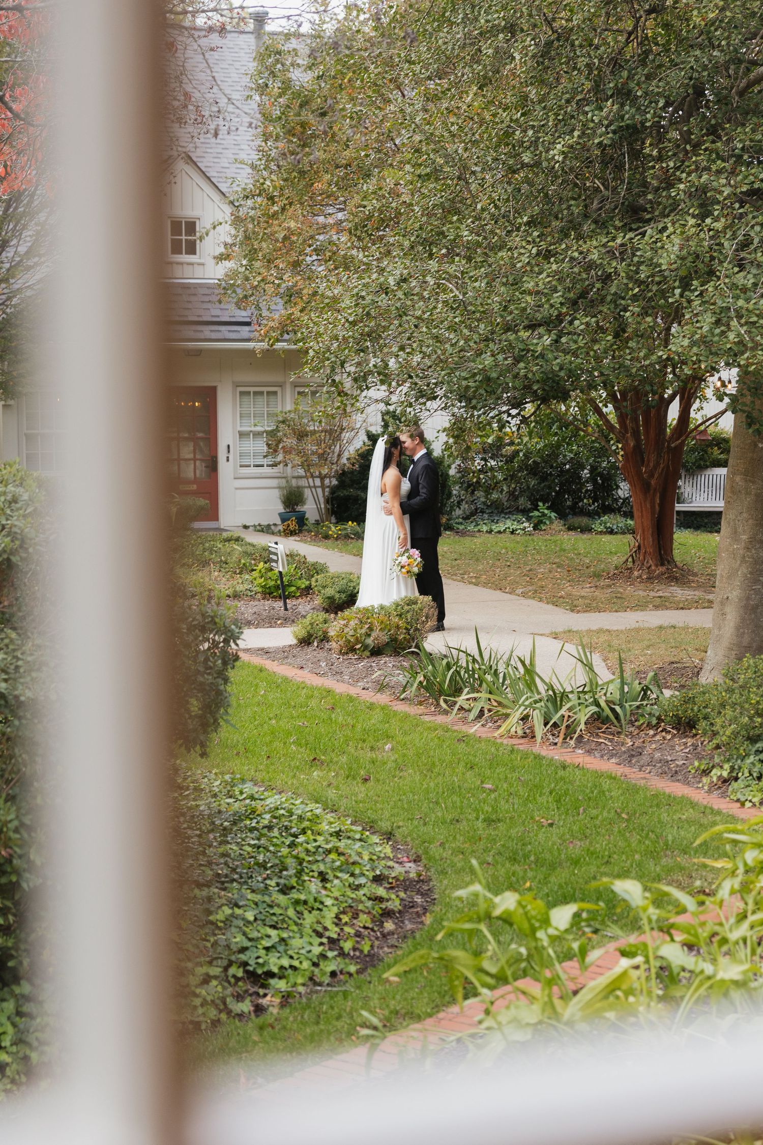 Bride and groom sharing an intimate moment in a lush garden, photographed through a window frame.