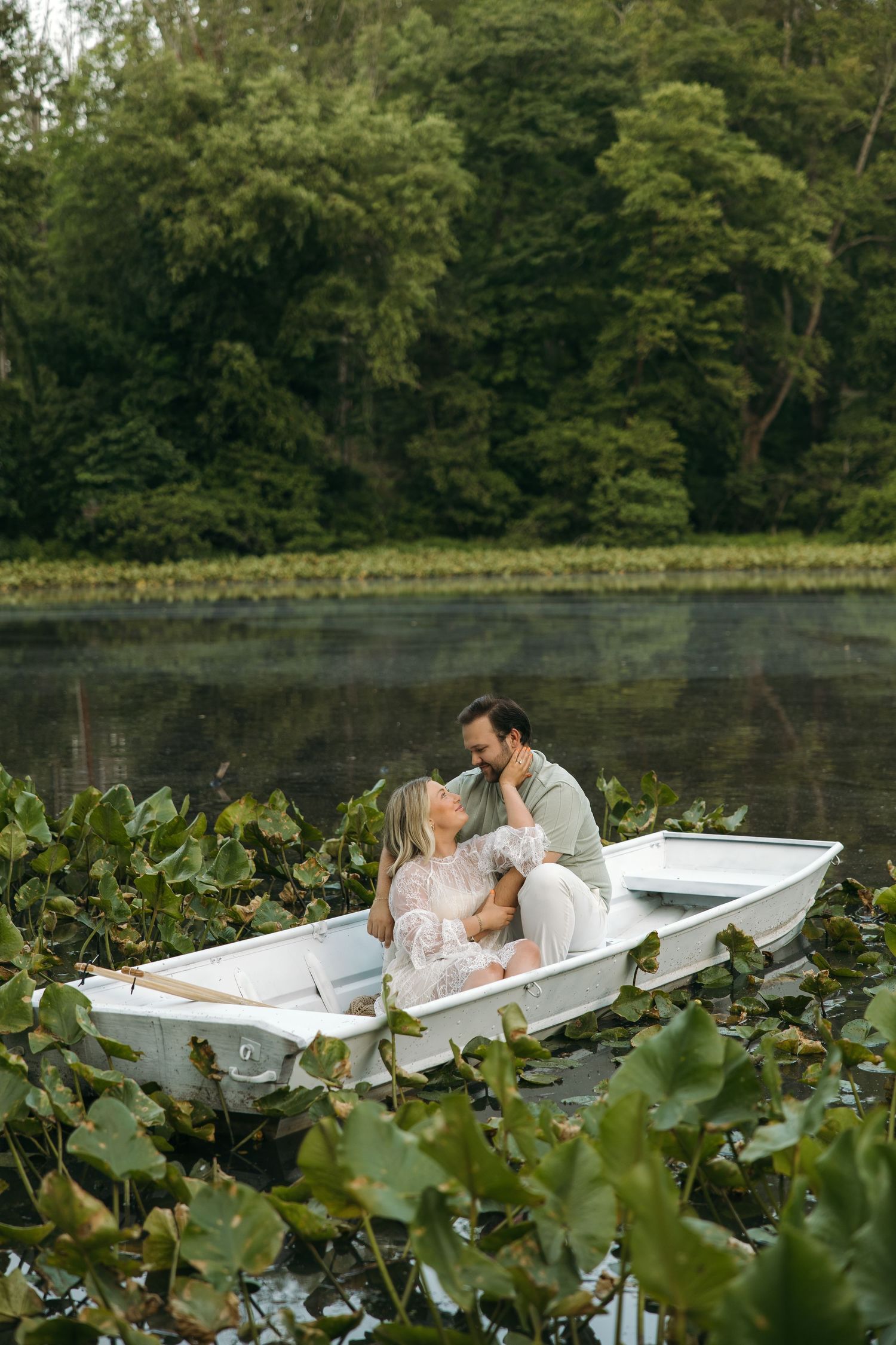A loving couple embraces and gazes at each other in a white rowboat surrounded by lily pads on a calm lake.