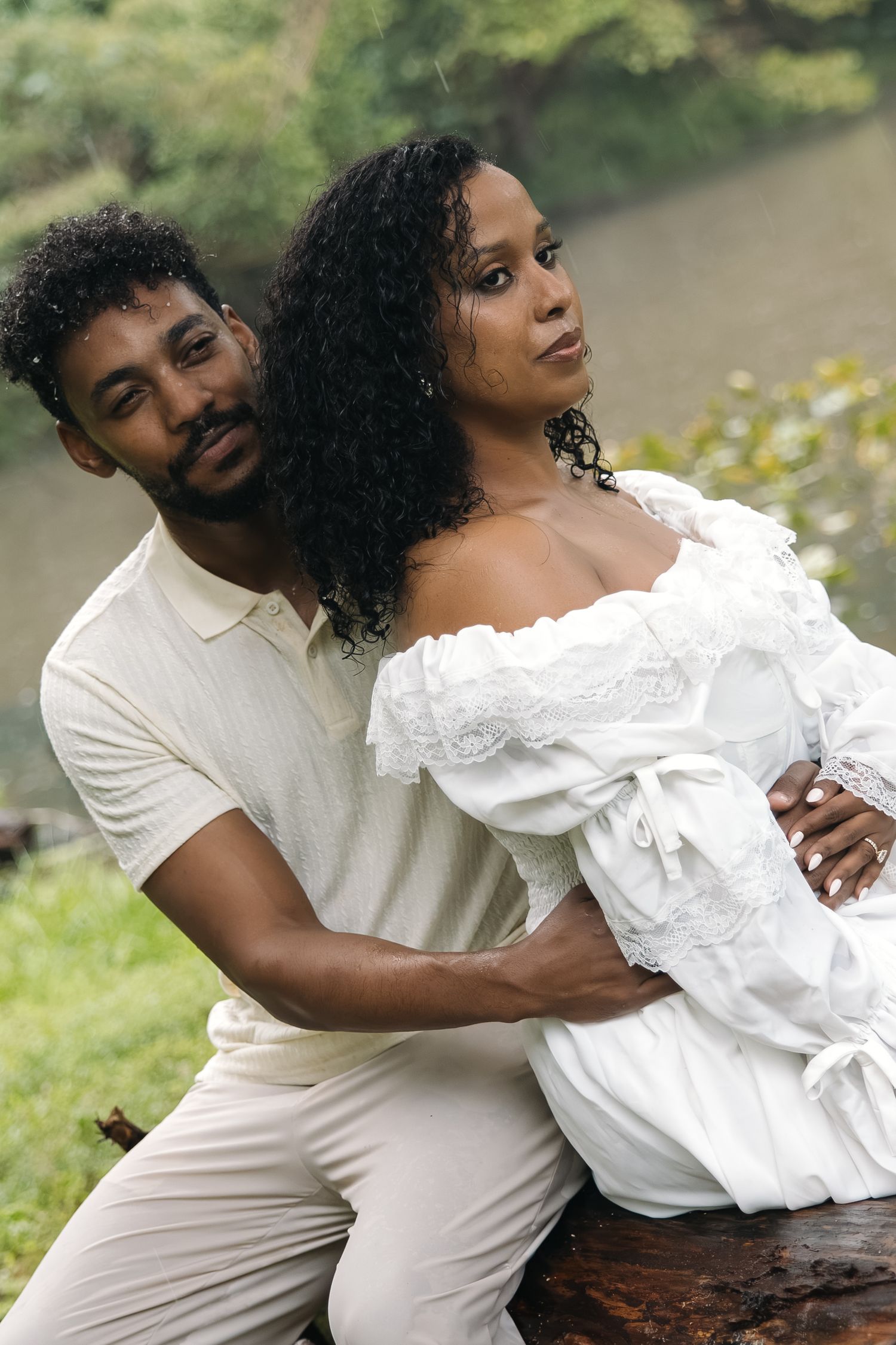 A couple in white outfits poses outdoors, the man embracing the woman from behind near a lush green background.