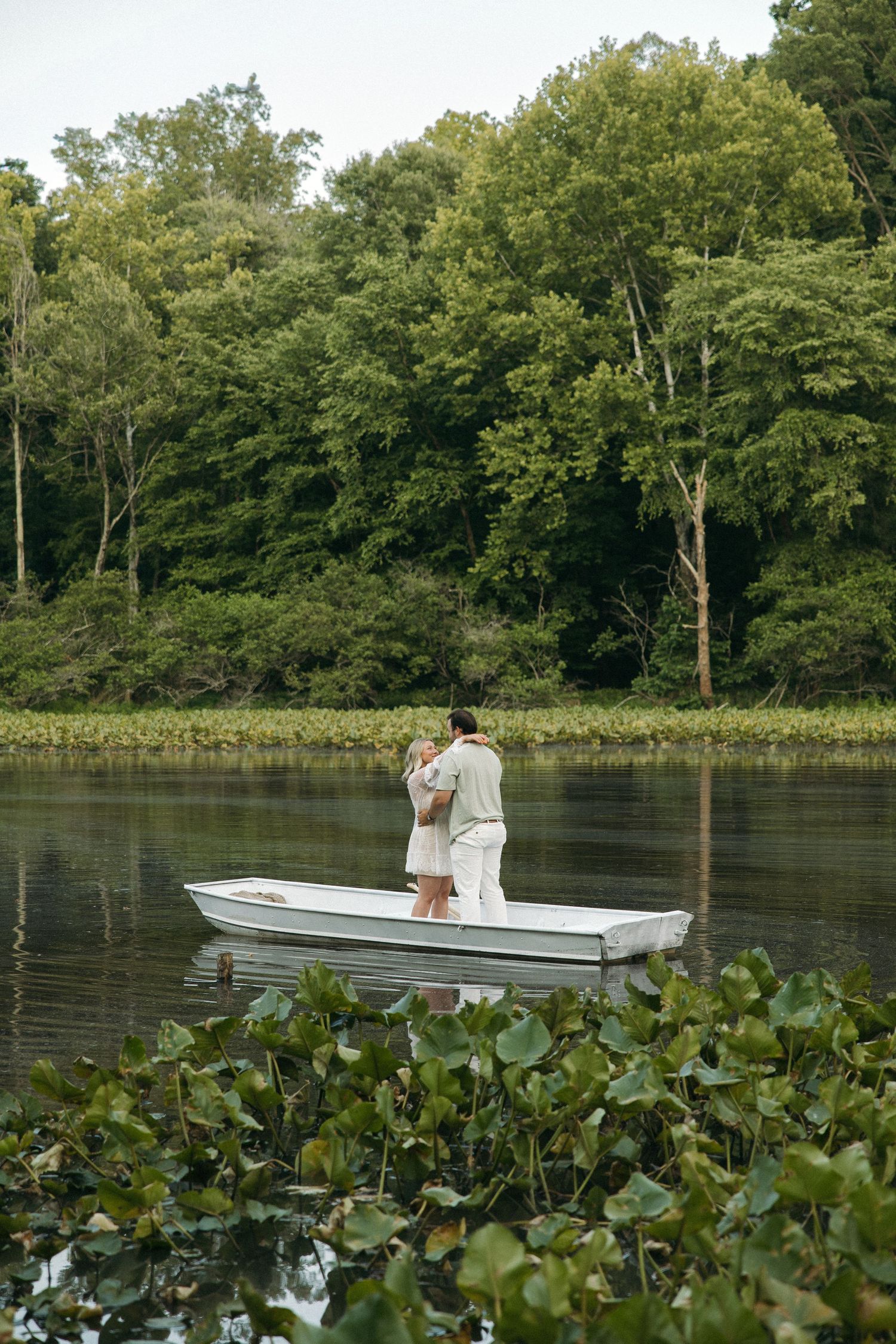 Couple standing and embracing in a white rowboat on a calm river with lush green trees.