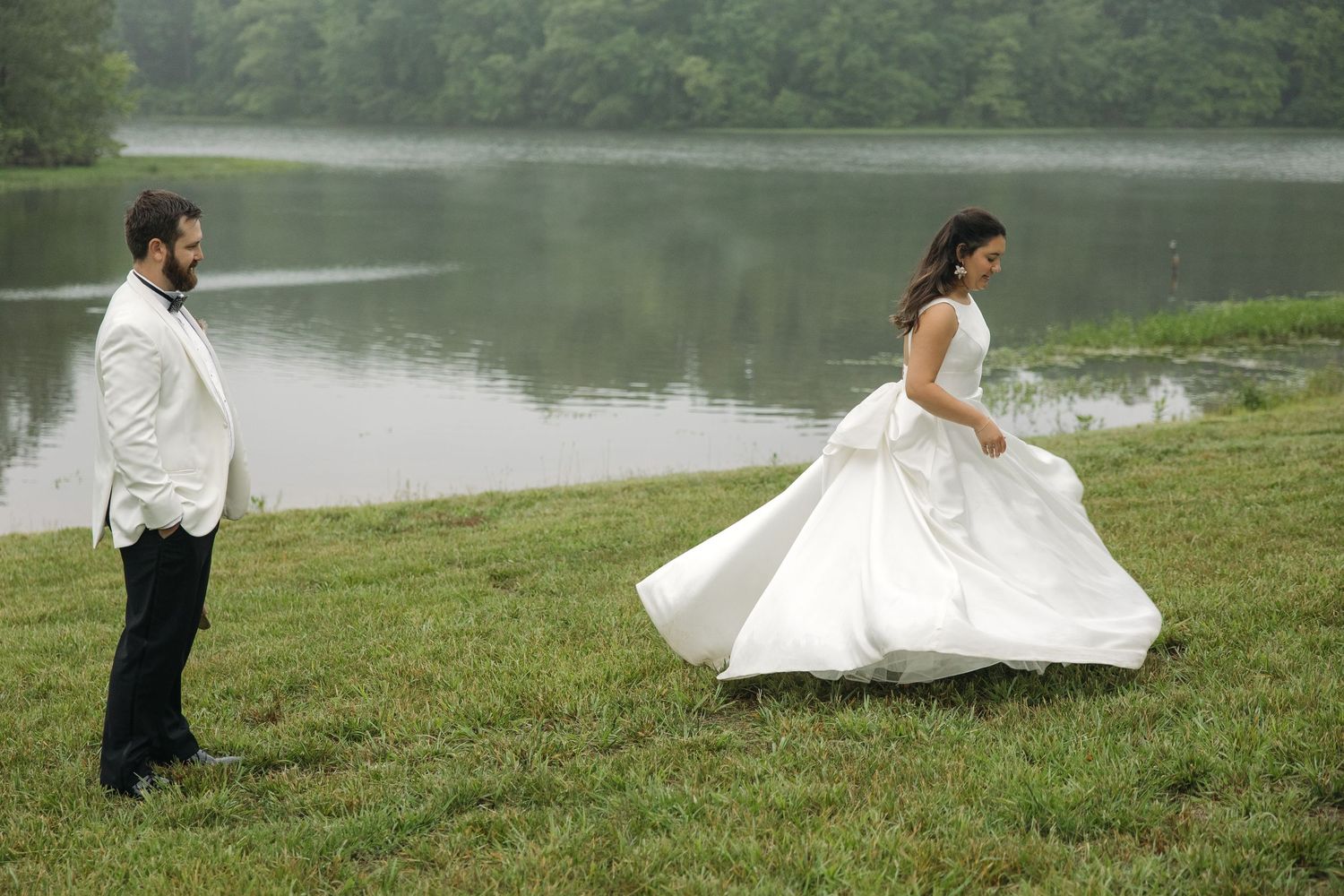 Bride in white ball gown twirls near lakeside as groom in white tuxedo watches on lush green grass.