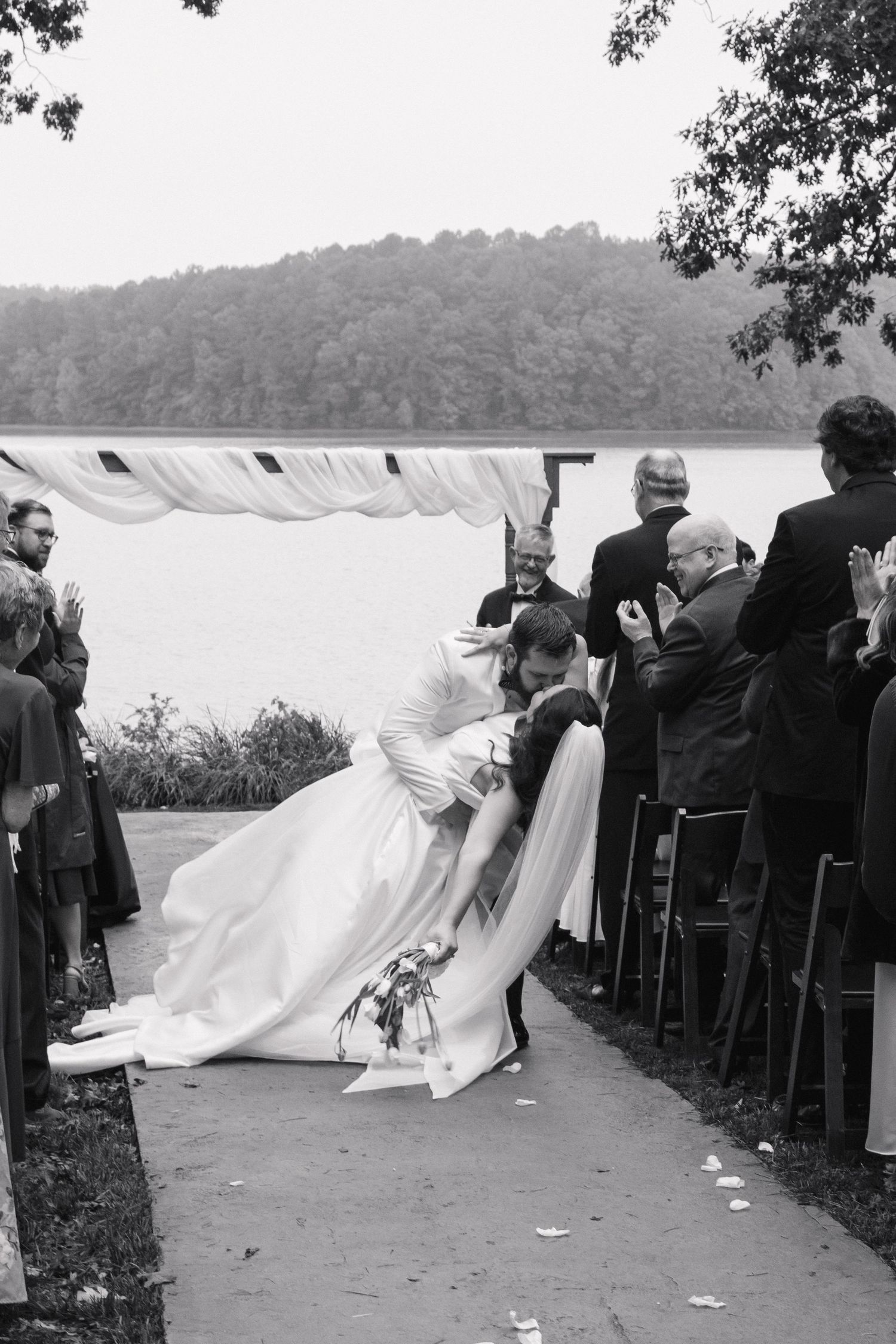 Black and white photo of groom dipping bride for a kiss at lakeside outdoor wedding ceremony with guests applauding.