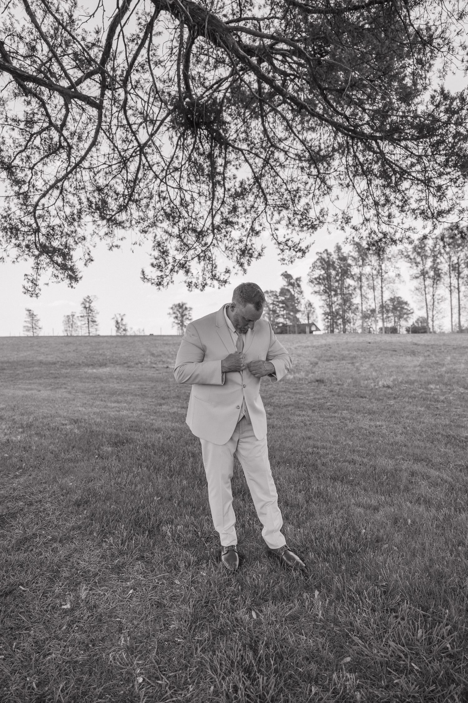 Groom in beige suit adjusting cufflinks outdoors under tree branches in black and white wedding photo.