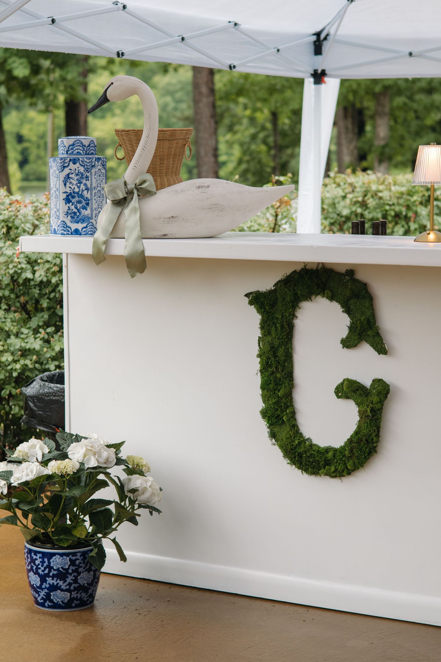 Elegant outdoor bar with moss letter G monogram, blue and white vase, and swan decor under white tent.