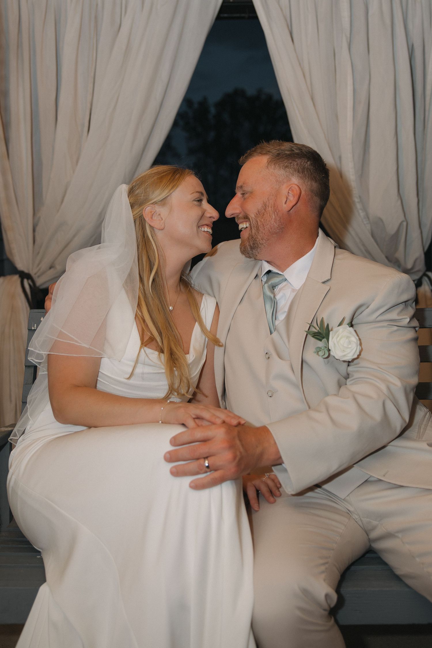 Bride and groom sit together laughing joyfully on a bench surrounded by elegant draped curtains at night.