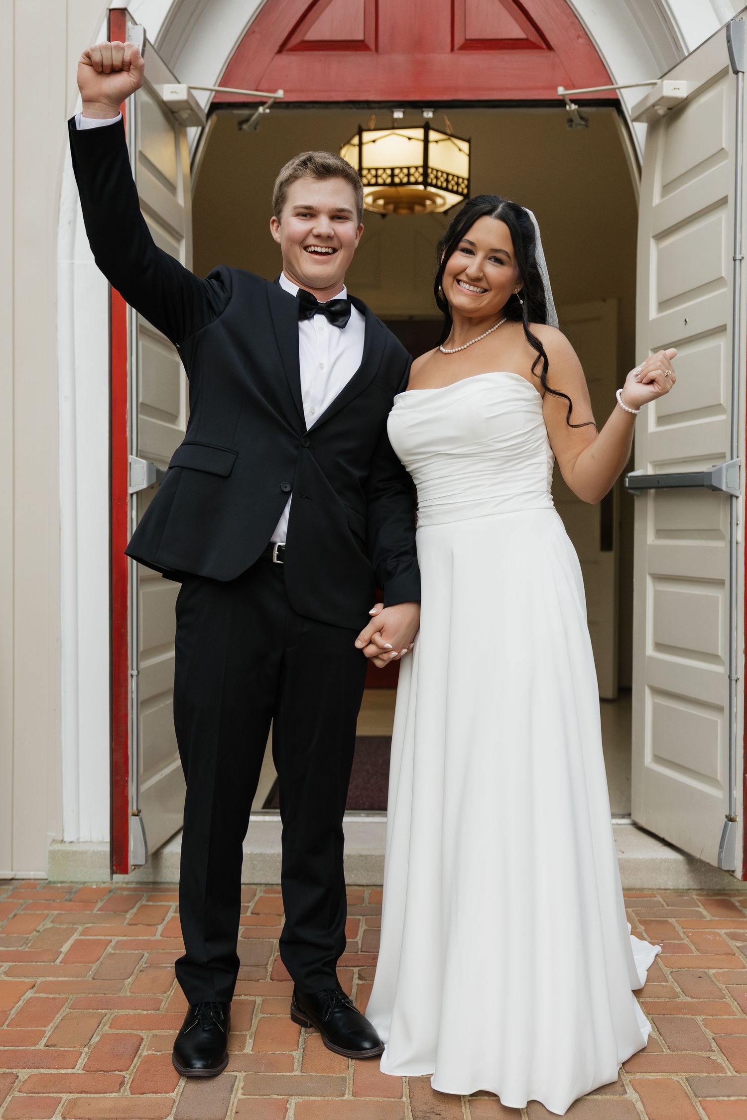 Joyful newlyweds celebrating outside church doors, groom raising fist and bride pointing in excitement.