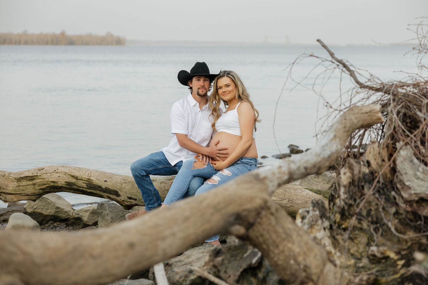 Happy couple posing on driftwood near a lake, man in black cowboy hat holding pregnant partners belly outdoors.