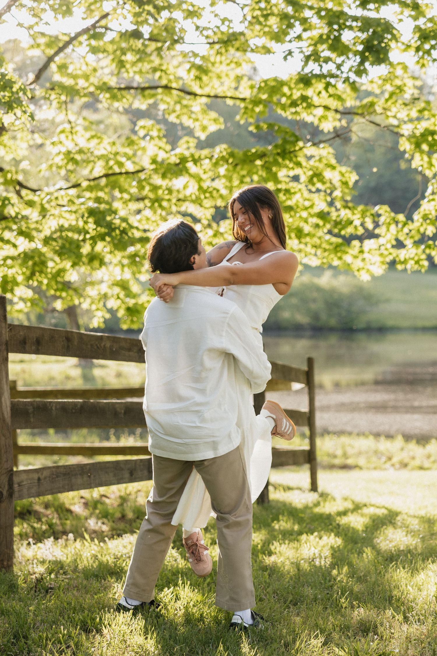 Man in white shirt joyfully lifting woman in white dress outdoors near a wooden fence in golden sunlight.