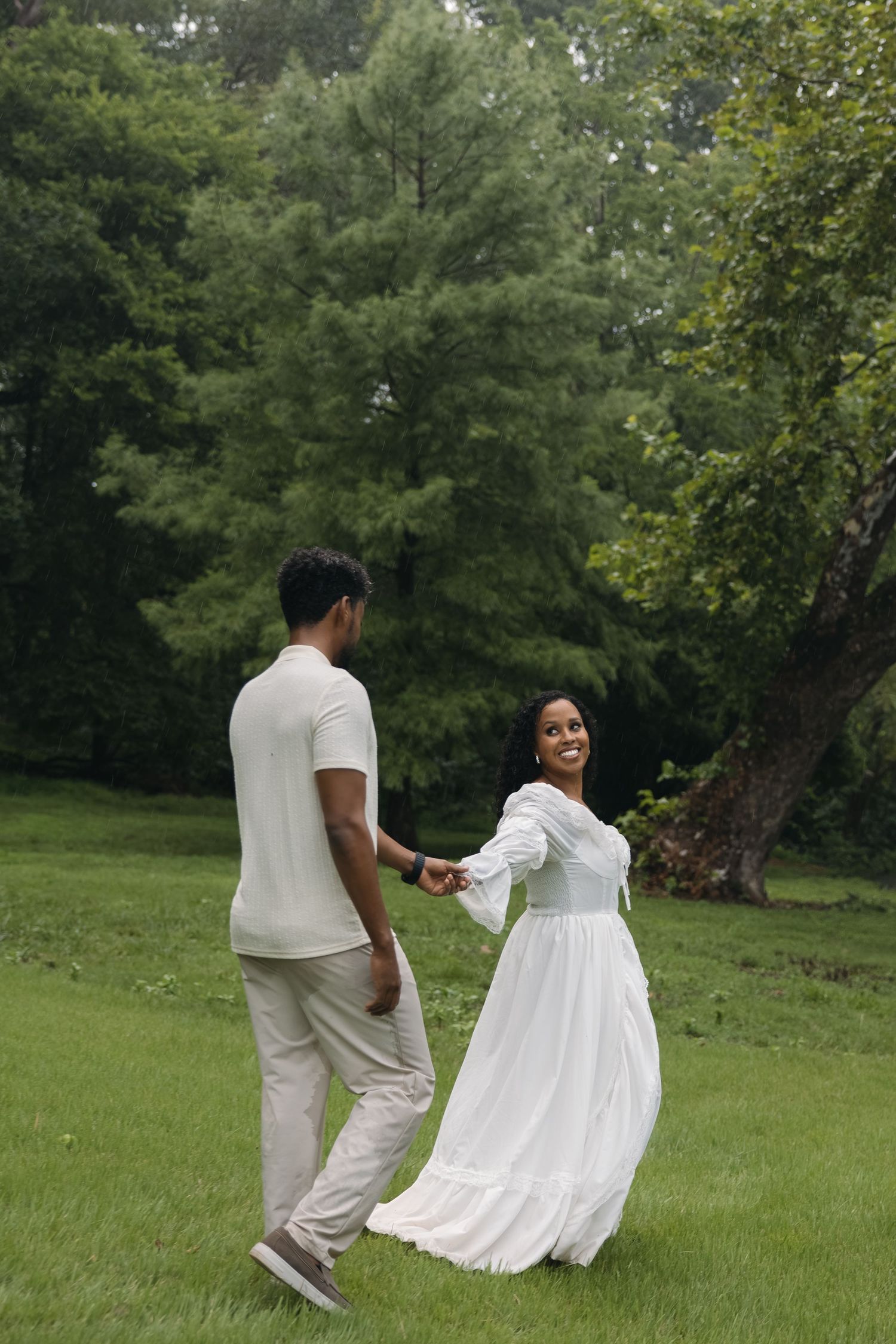 A couple holds hands while walking on green grass, the woman wearing a flowing white dress, surrounded by lush trees.