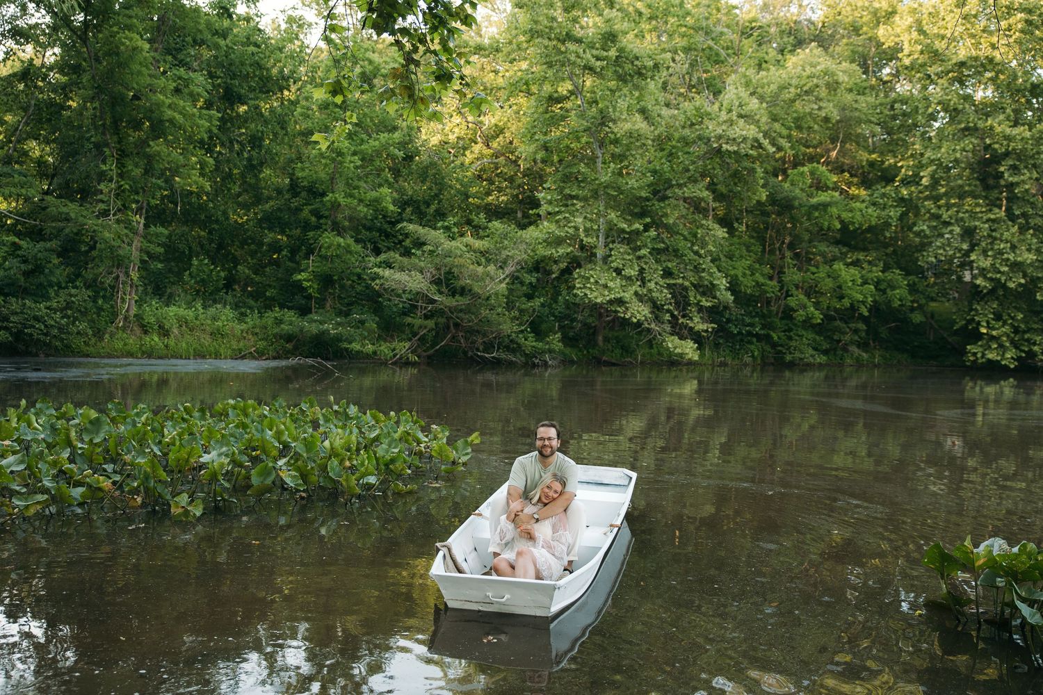 A couple shares a peaceful moment in a small white boat floating on a calm tree-lined river.