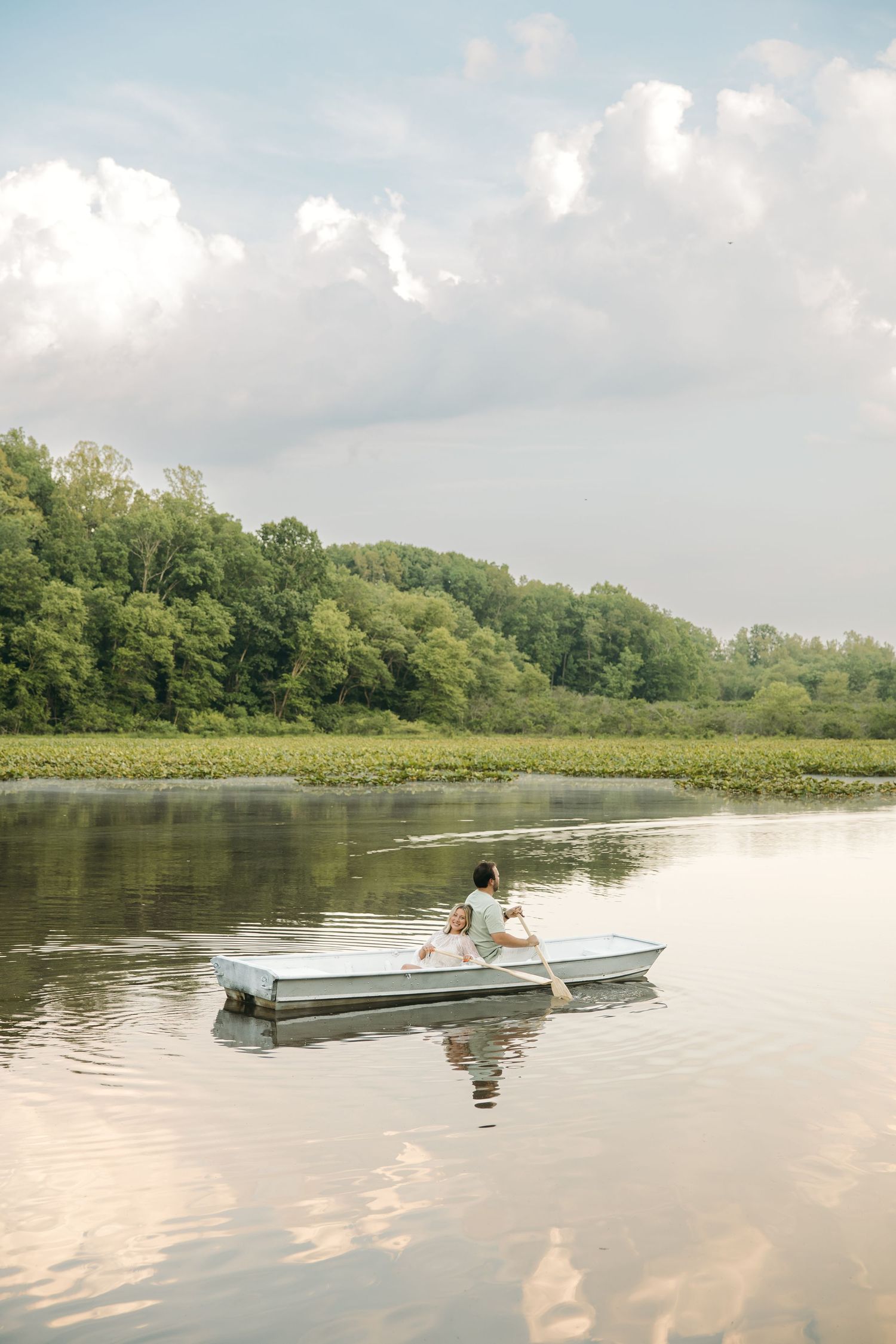 Person sits alone in a white rowboat on a glassy lake reflecting clouds and lush green forest trees.