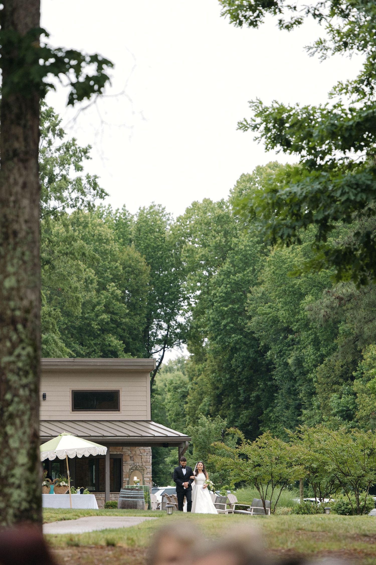 Bride and escort walking toward outdoor wedding ceremony venue surrounded by lush green trees.