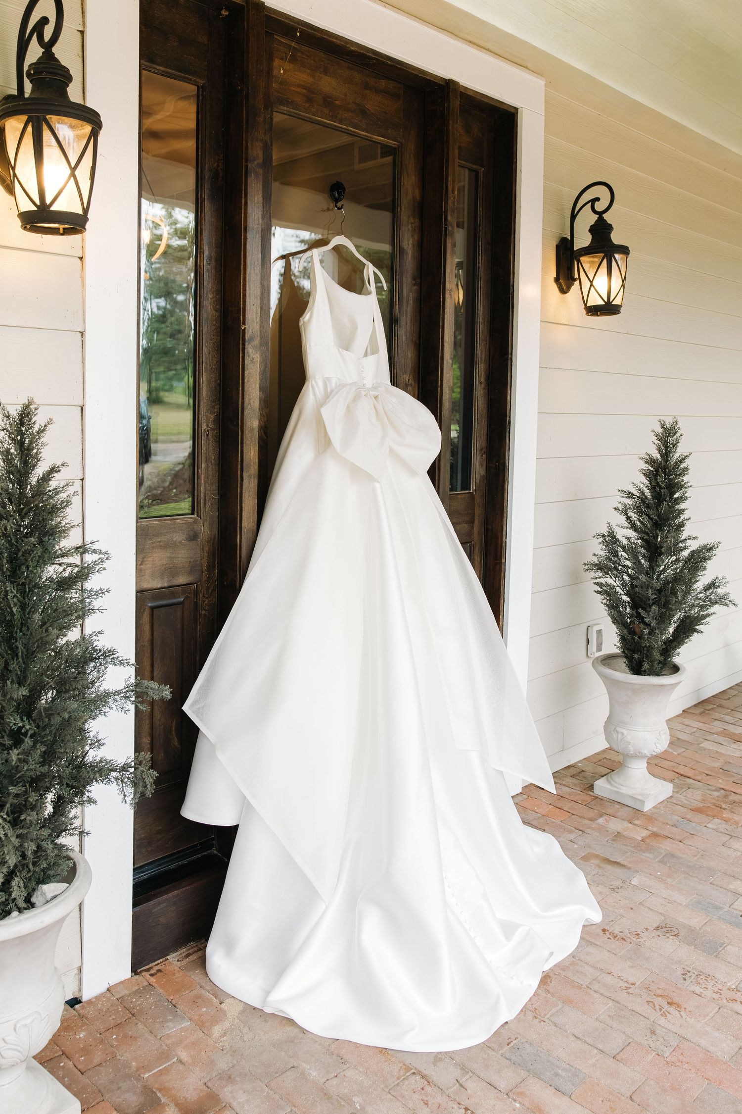 Stunning white satin wedding dress with large bow detail hanging on a brick porch with wooden double doors.