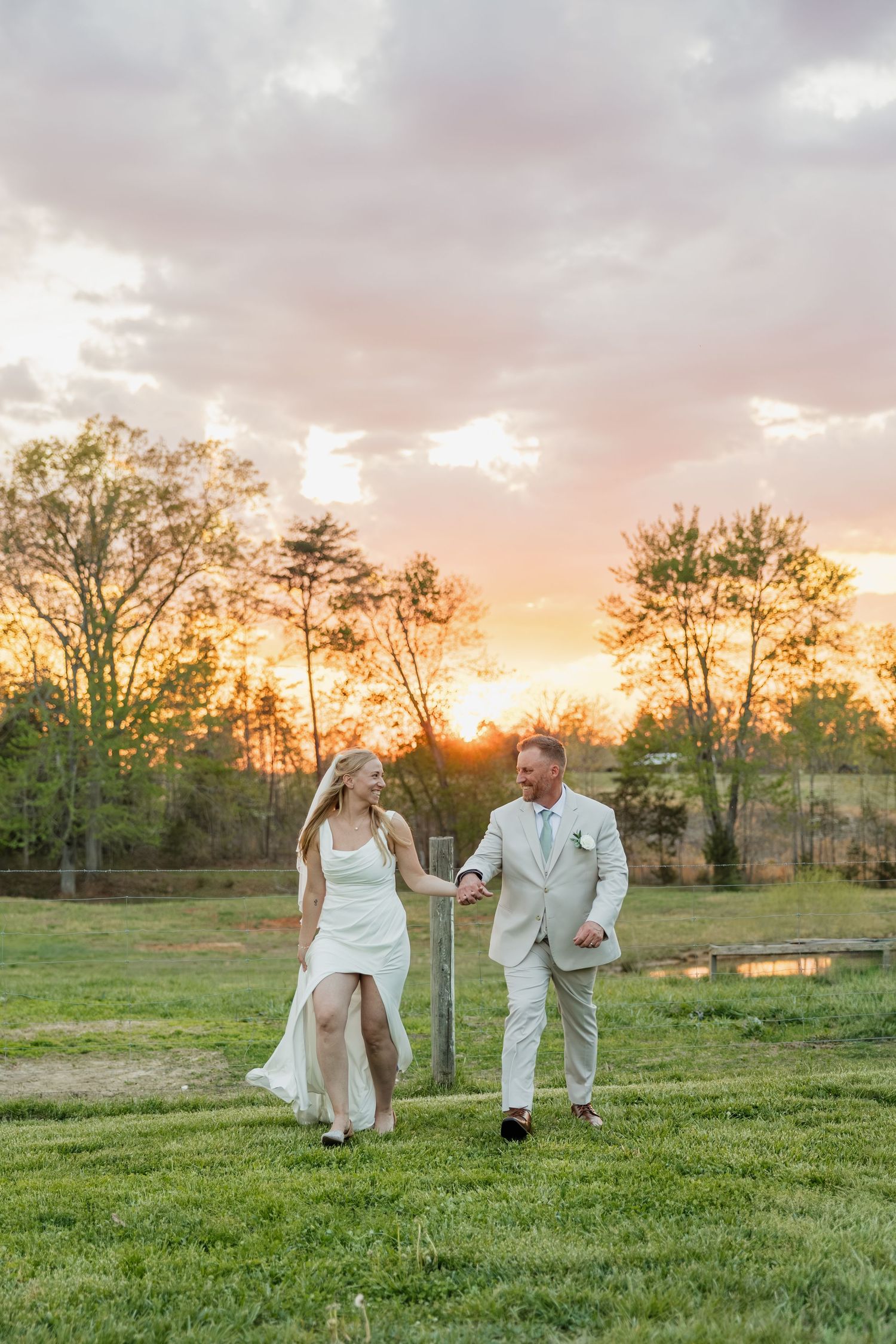 Smiling bride and groom holding hands and walking joyfully across a green field at sunset.