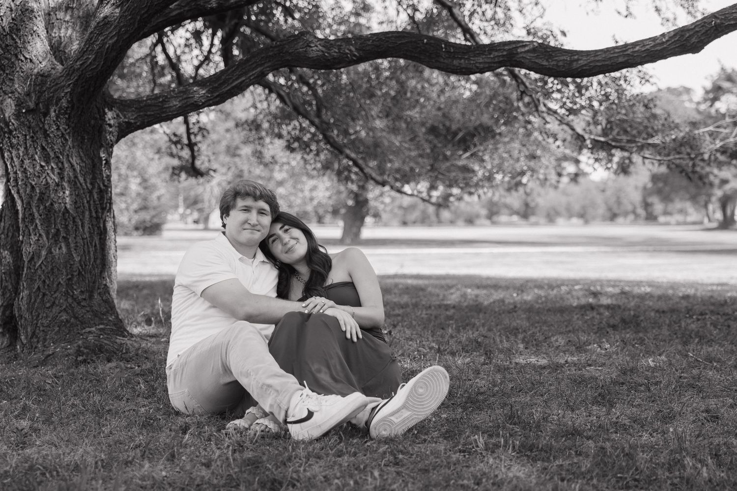 Black and white photo of a couple sitting together under a tree in a park, smiling and embracing.
