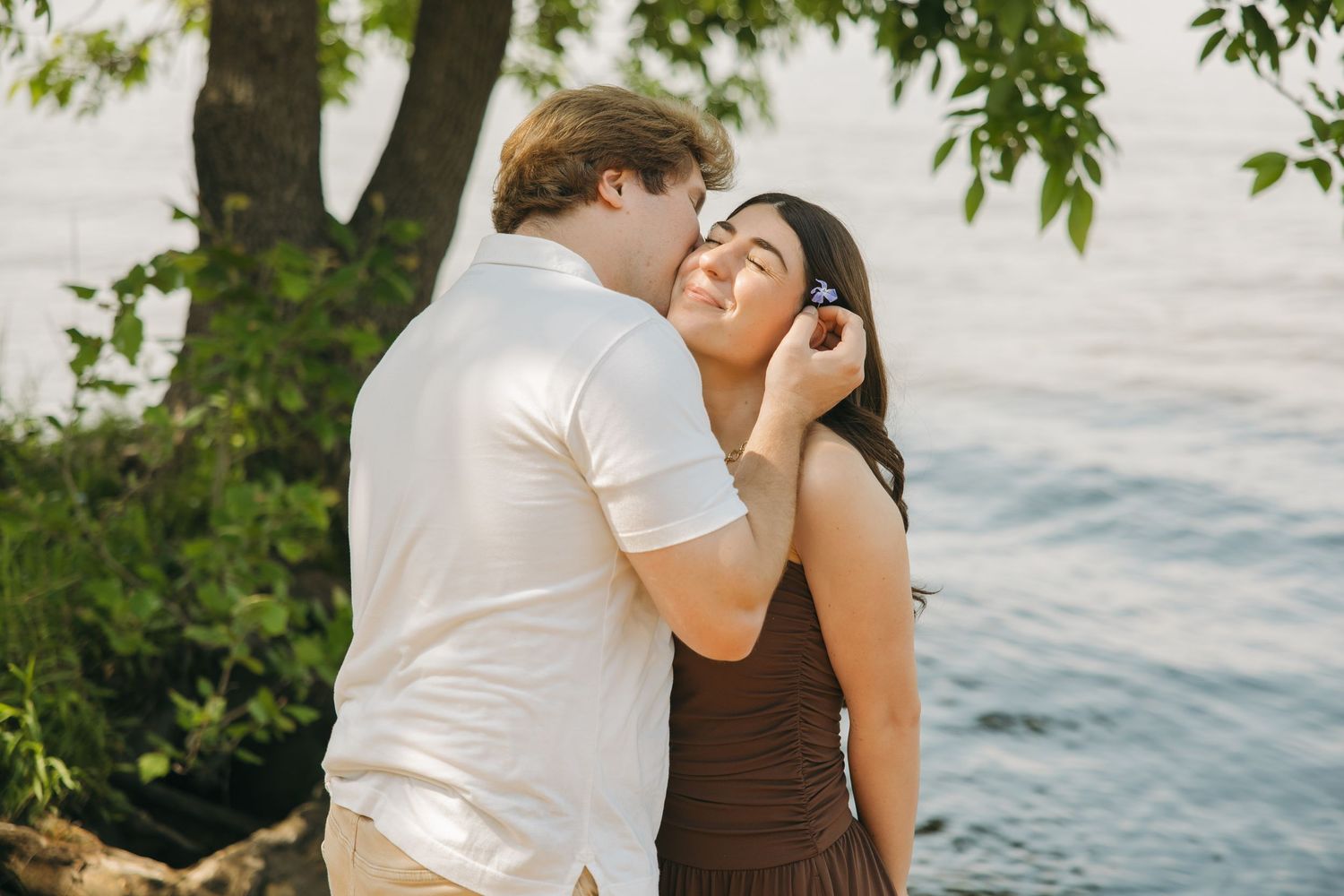 Man tenderly kissing his girlfriends cheek near a sunlit lakeside setting.