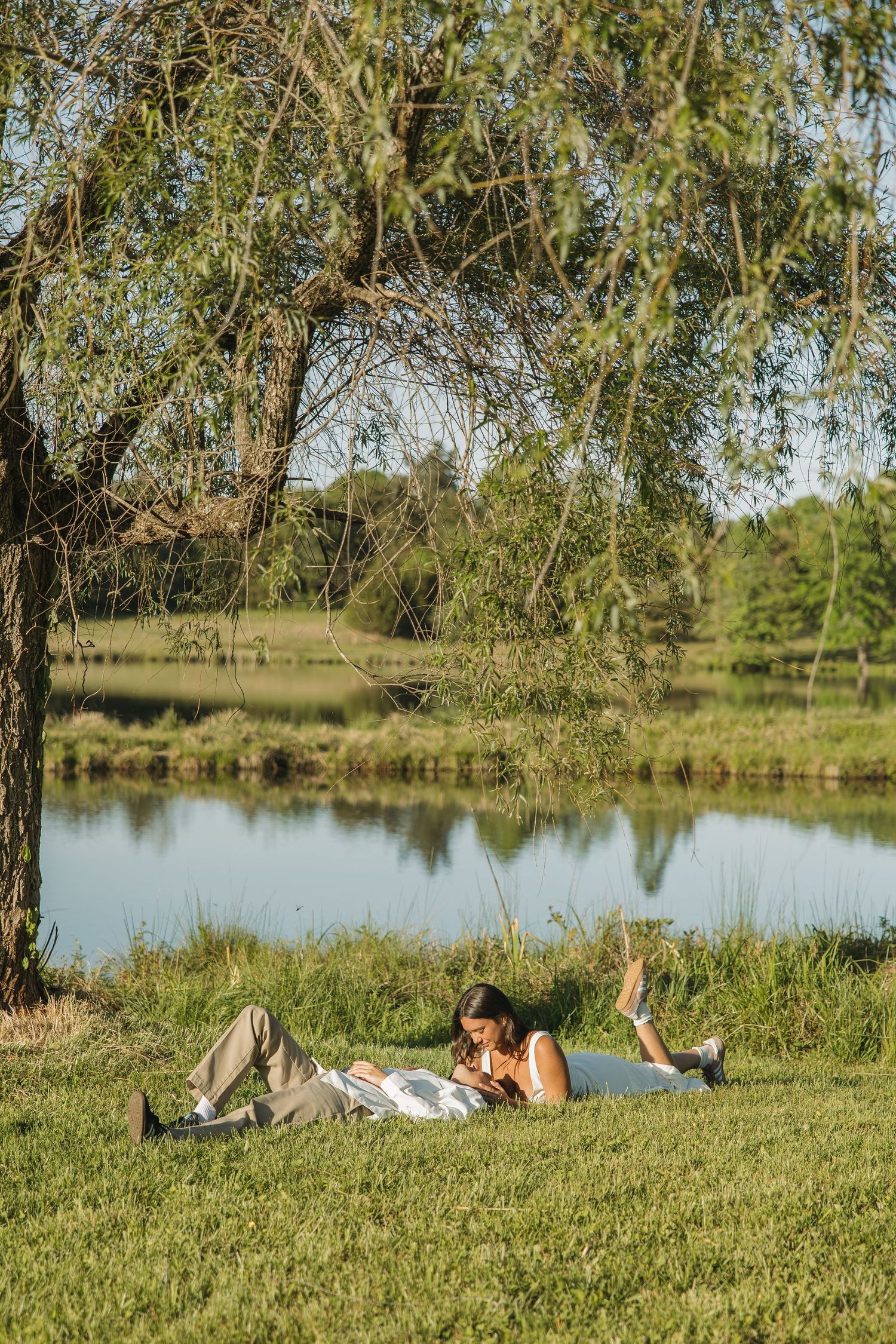Couple lying on grass under a willow tree by a calm lake, relaxing together on a sunny afternoon.