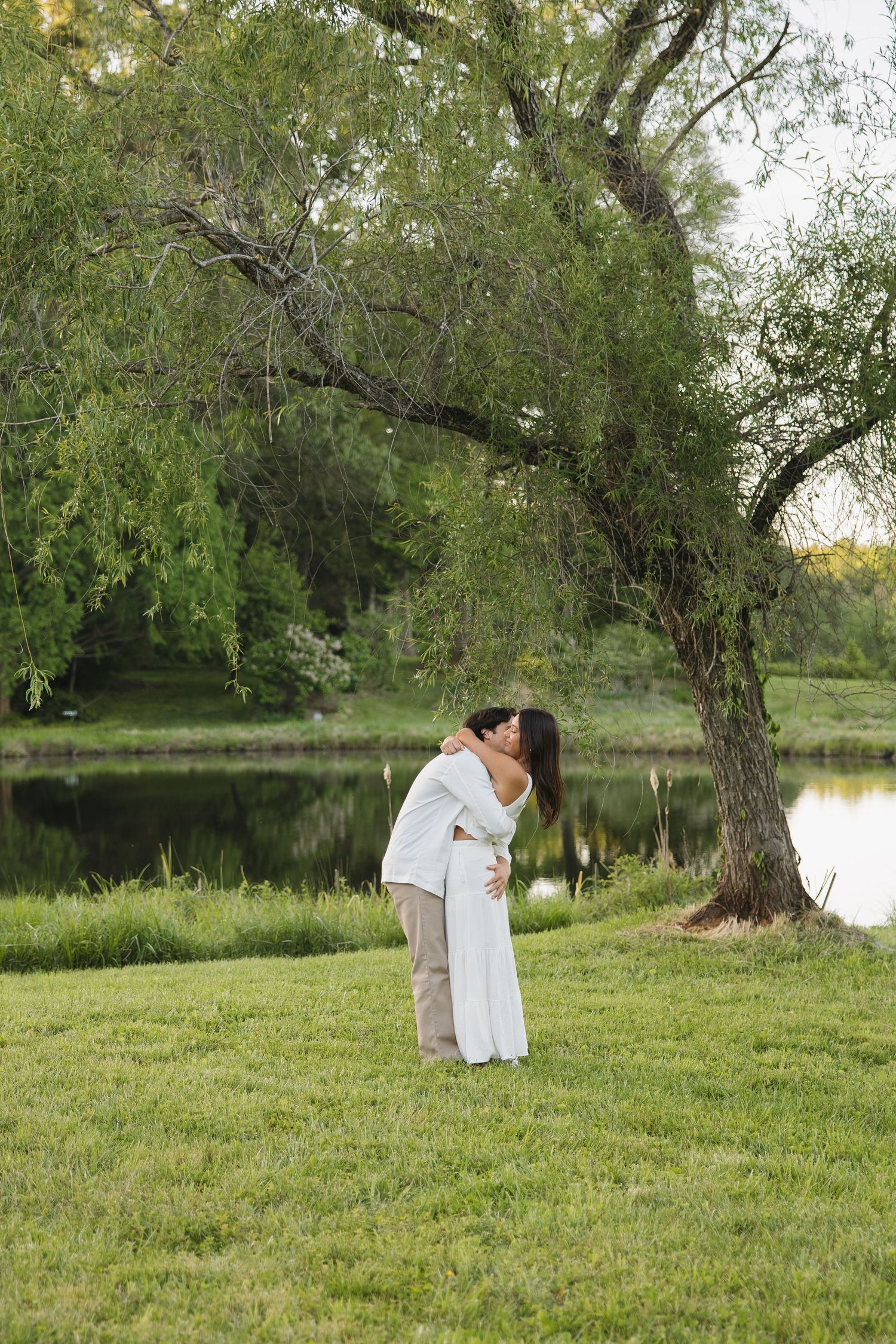 Couple sharing a romantic kiss under a willow tree beside a calm reflective pond at dusk.