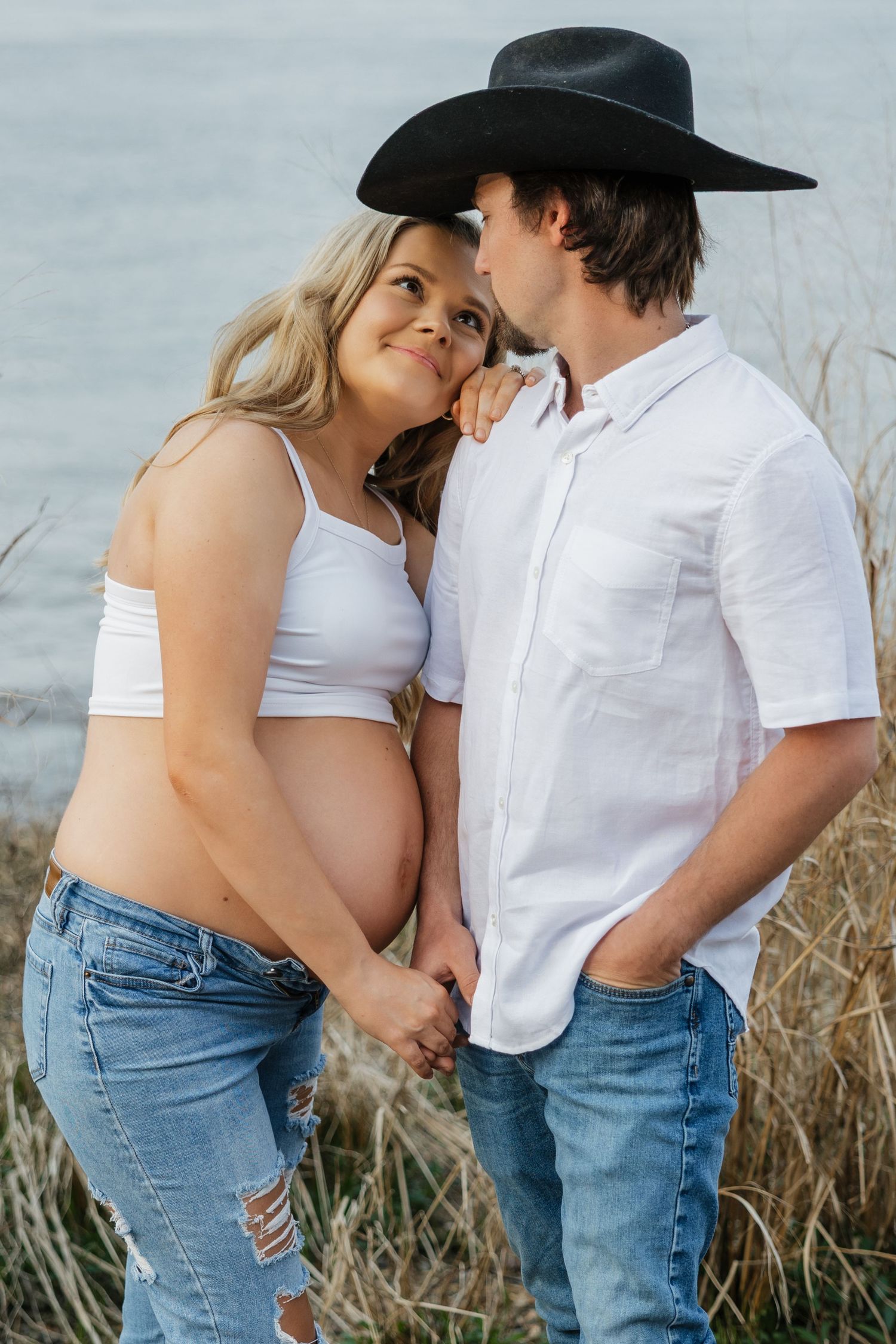 Pregnant woman in white crop top holds hands with man in black cowboy hat near lake with tall grass background.