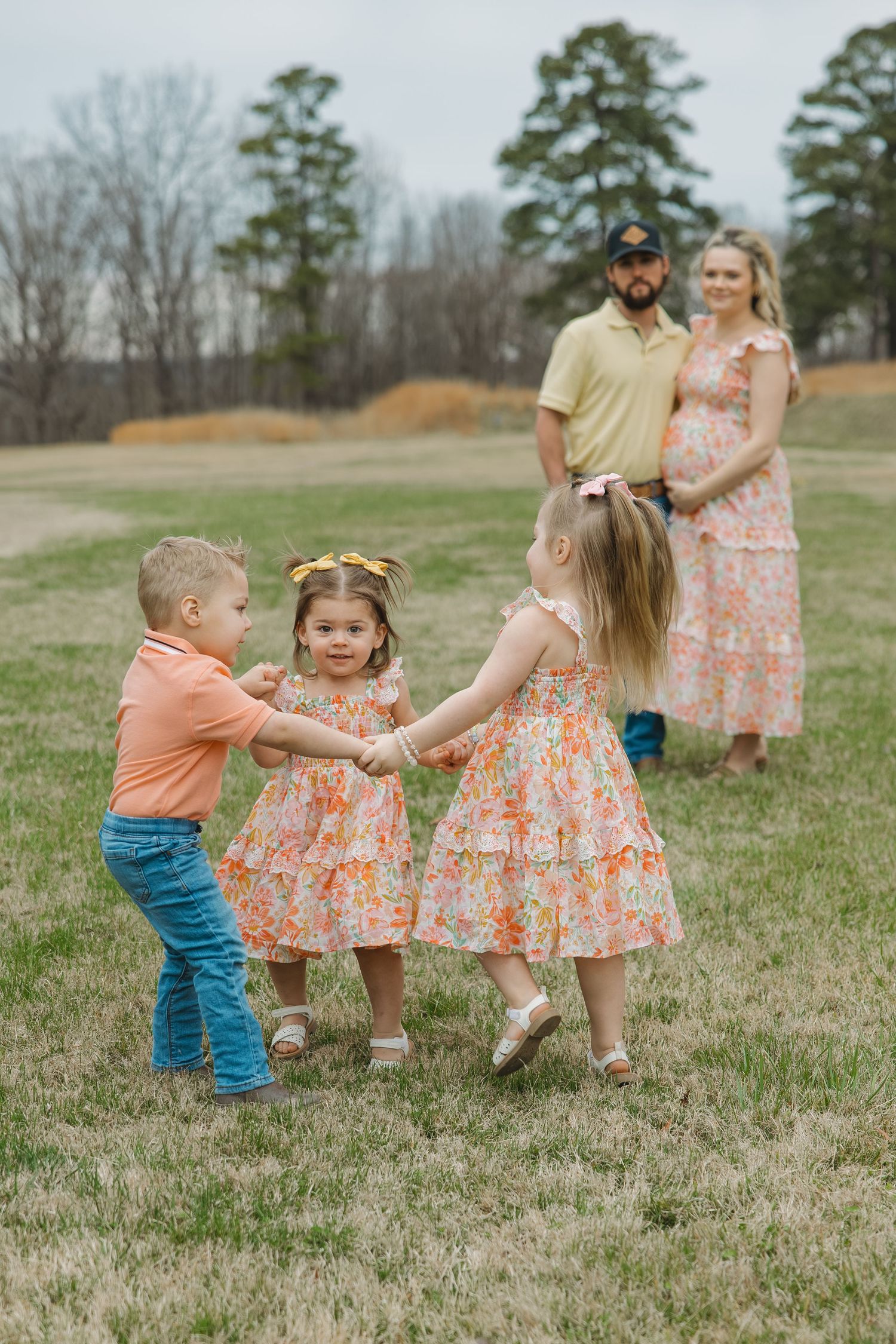 Young family with pregnant mother watches three children in matching outfits hold hands in a circle on a grassy field.