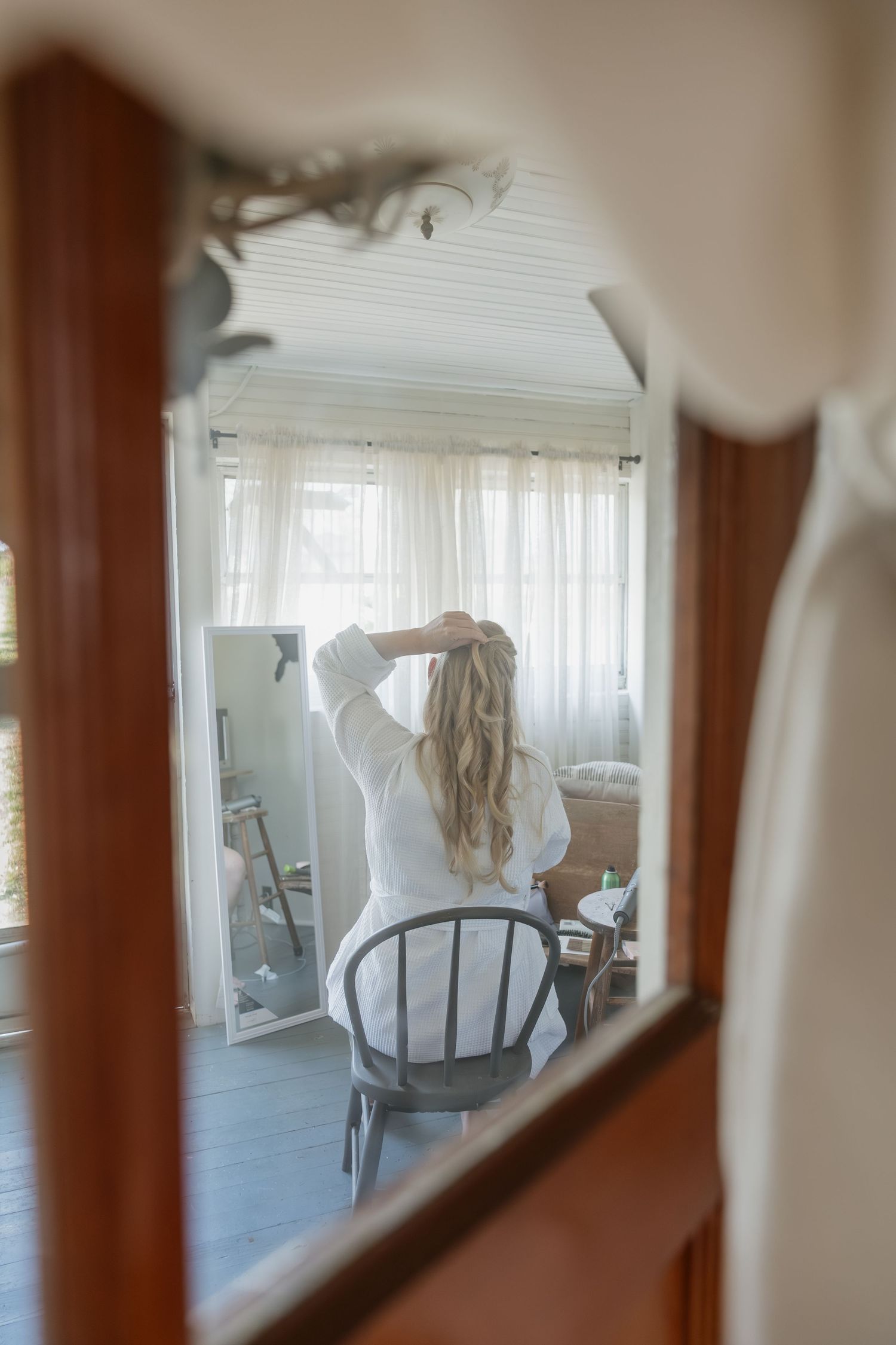 Bride in white robe styling her long blonde hair reflected in an ornate vintage mirror before wedding.