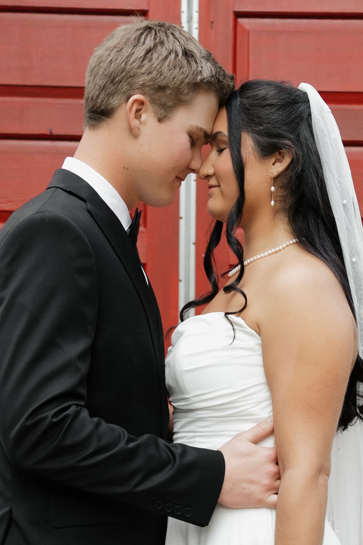 Bride and groom share an intimate forehead touch moment in front of a red door on their wedding day.