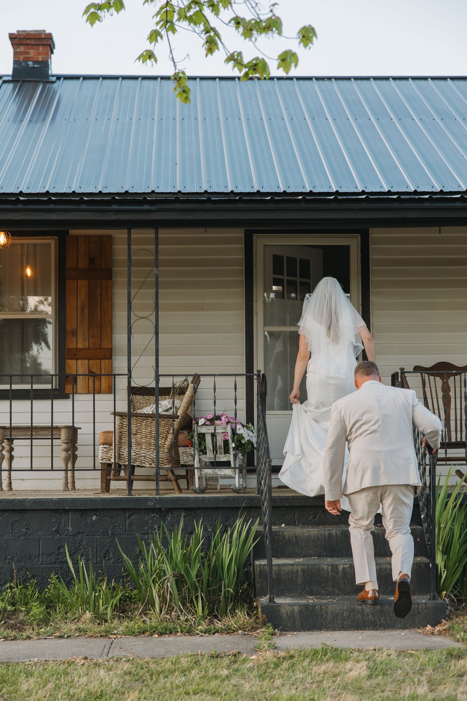 Bride and groom walk up porch steps of a charming white farmhouse after their wedding ceremony.