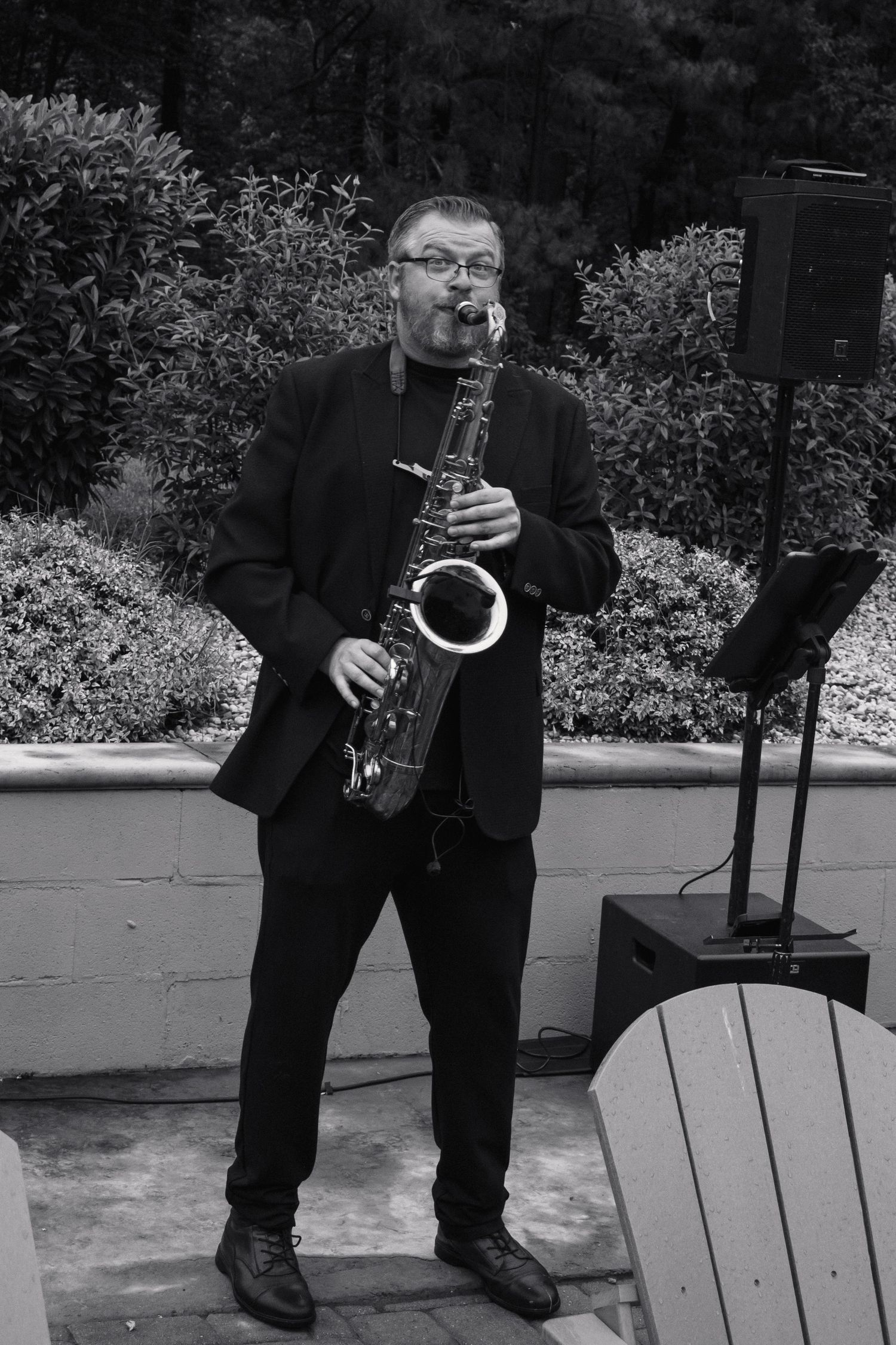 Black and white photo of saxophonist performing outdoors at wedding reception surrounded by lush greenery.