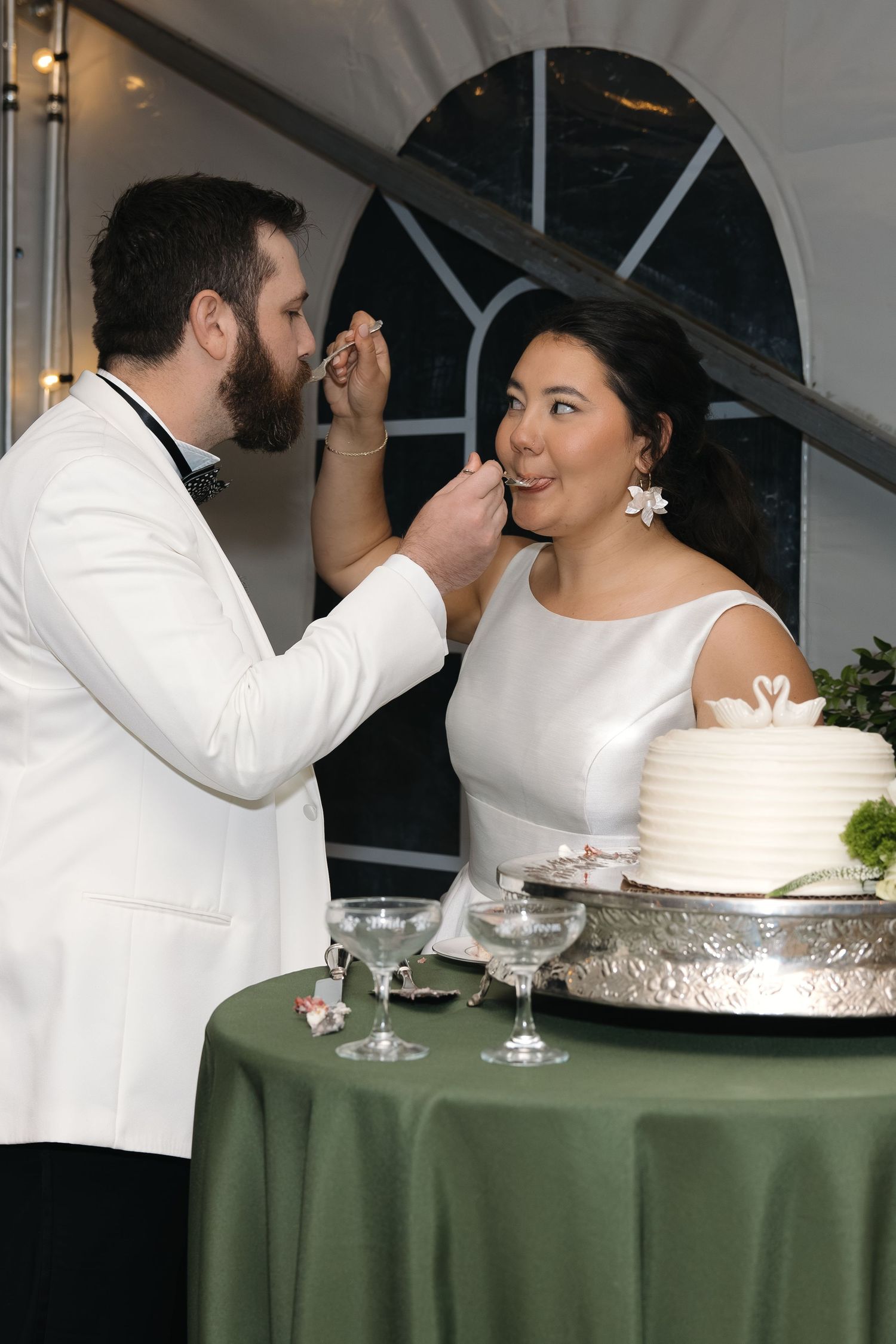 Groom in white jacket feeds wedding cake to smiling bride at elegant reception celebration.