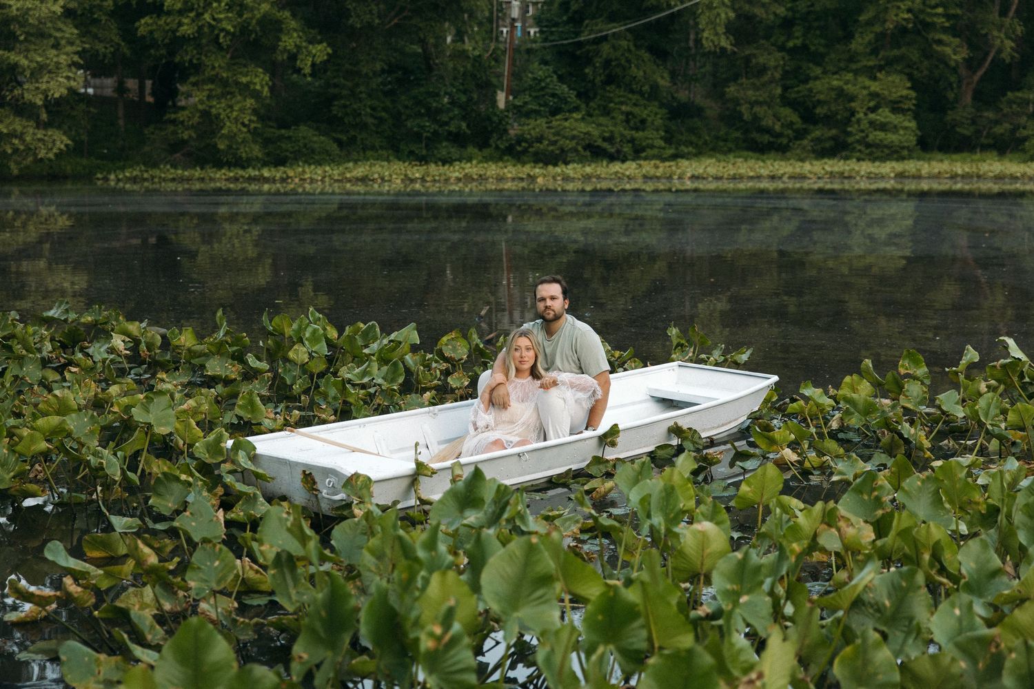 A happy couple poses together in a white rowboat surrounded by lily pads on a calm, tree-lined lake.