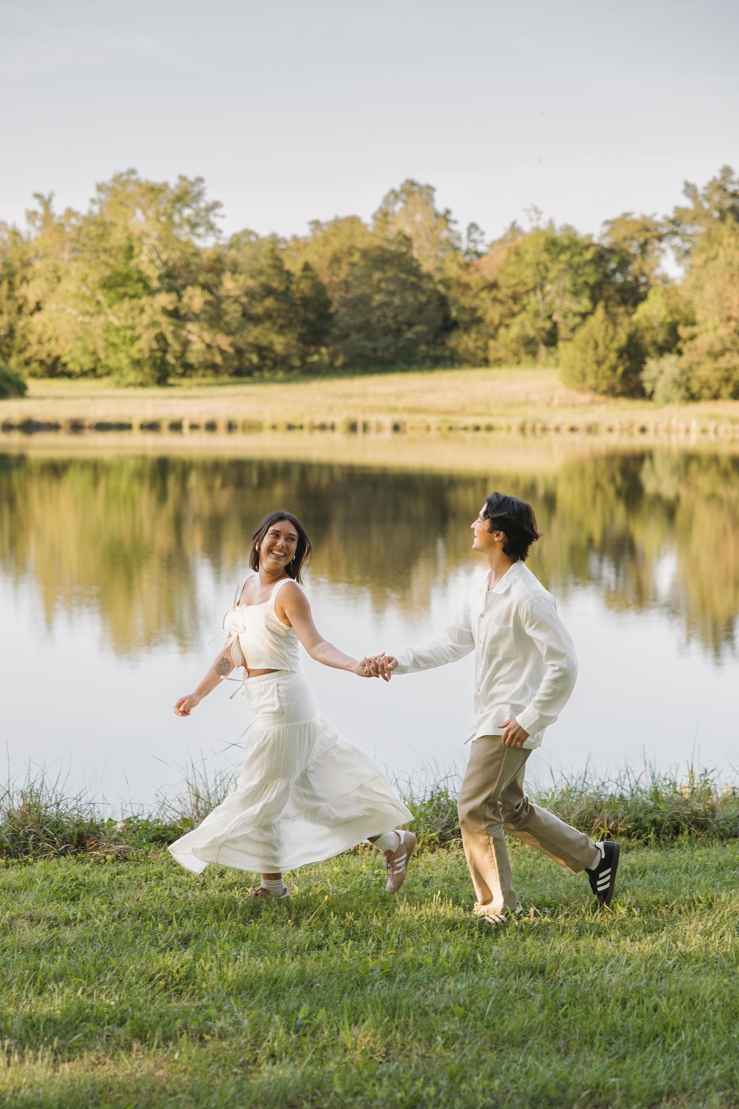 Couple running and holding hands along a scenic lakeside meadow on a sunny day.