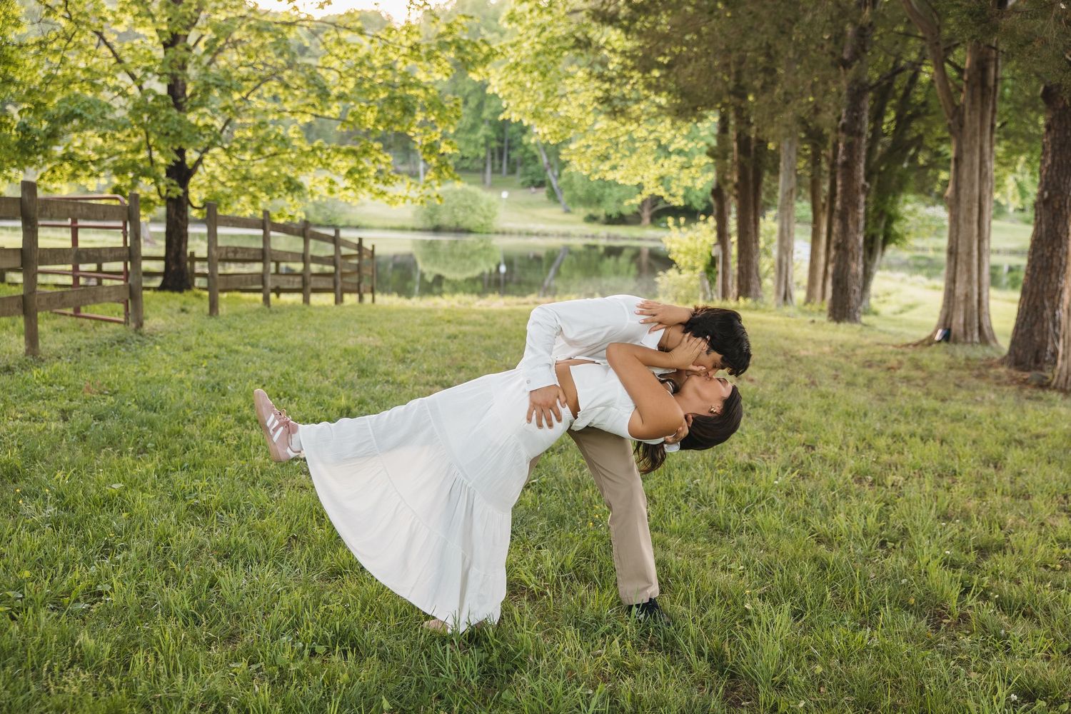 A couple shares a romantic dip kiss on a green lawn with trees and a lake behind them.