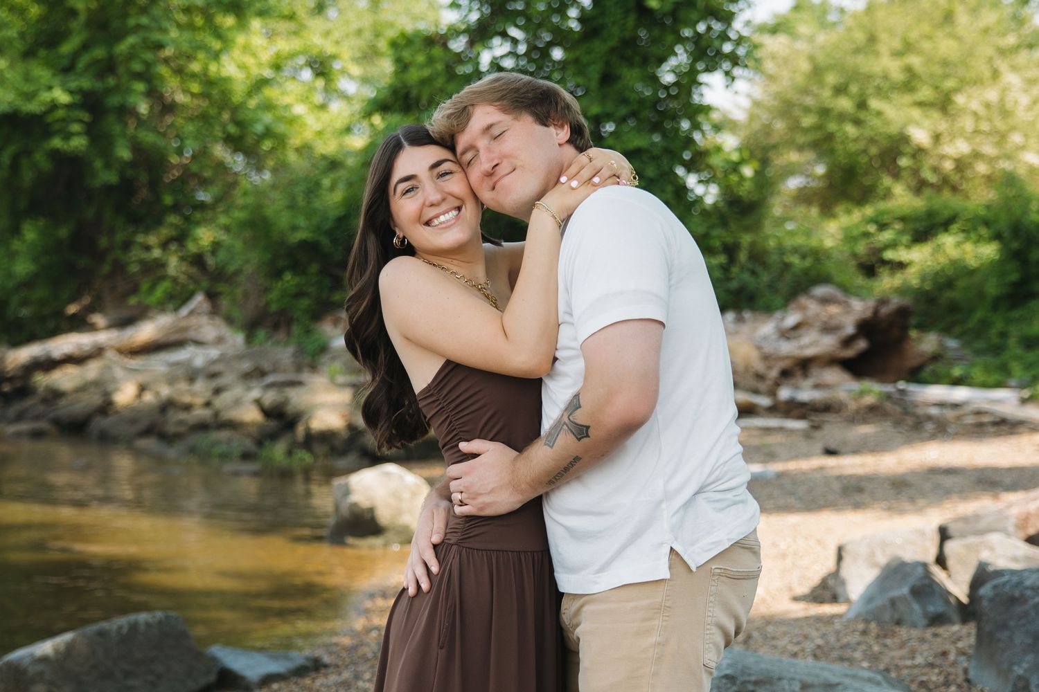 A smiling couple embraces warmly on a rocky riverbank with lush green trees in the background.