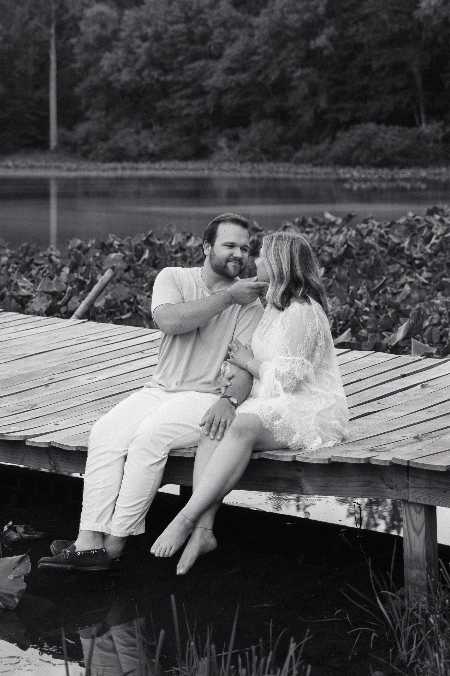 Black and white photo of a couple sitting intimately on a wooden dock by a lake, smiling and touching faces.