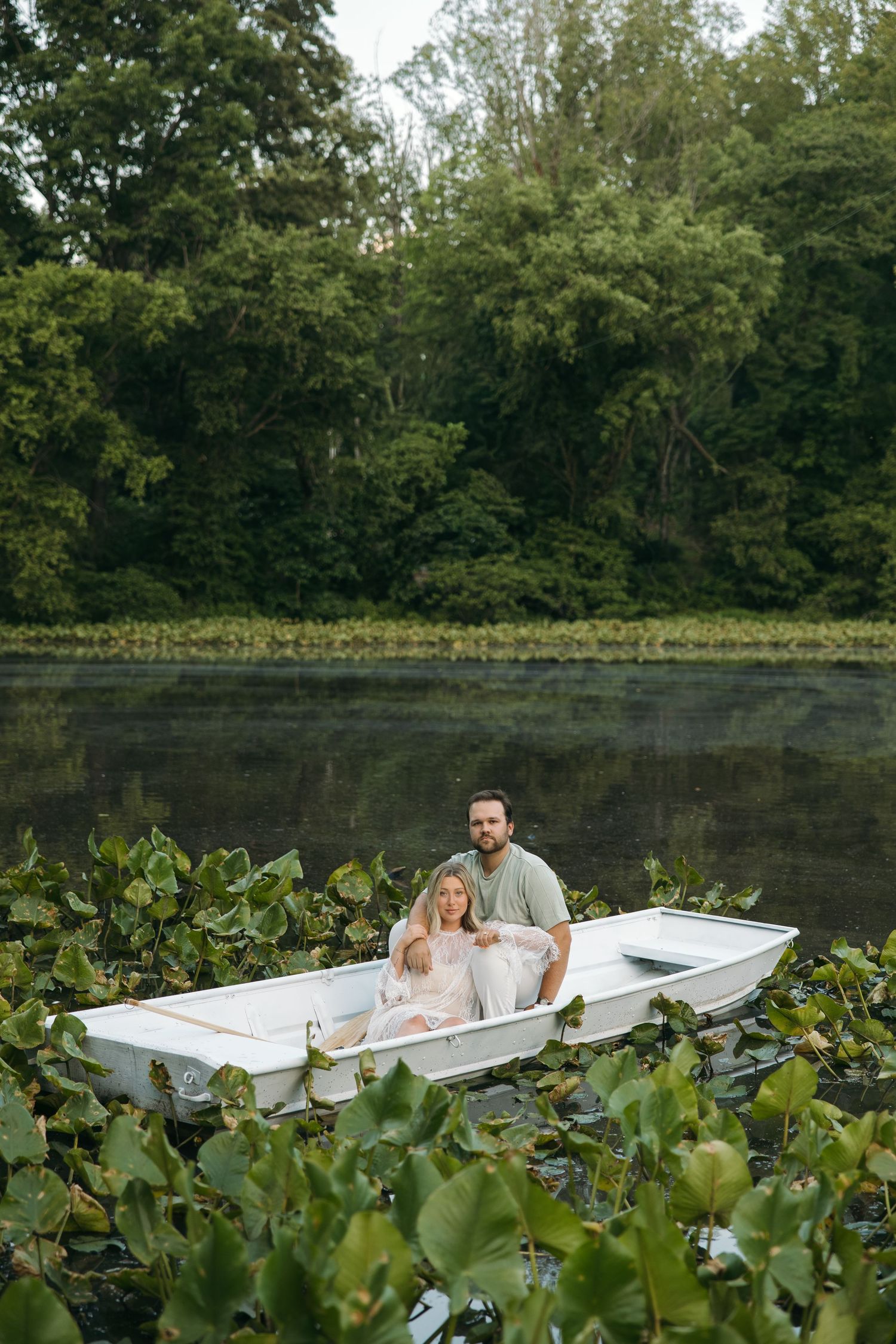 Couple sitting together in a white rowboat surrounded by lily pads on a serene lake with lush green trees.