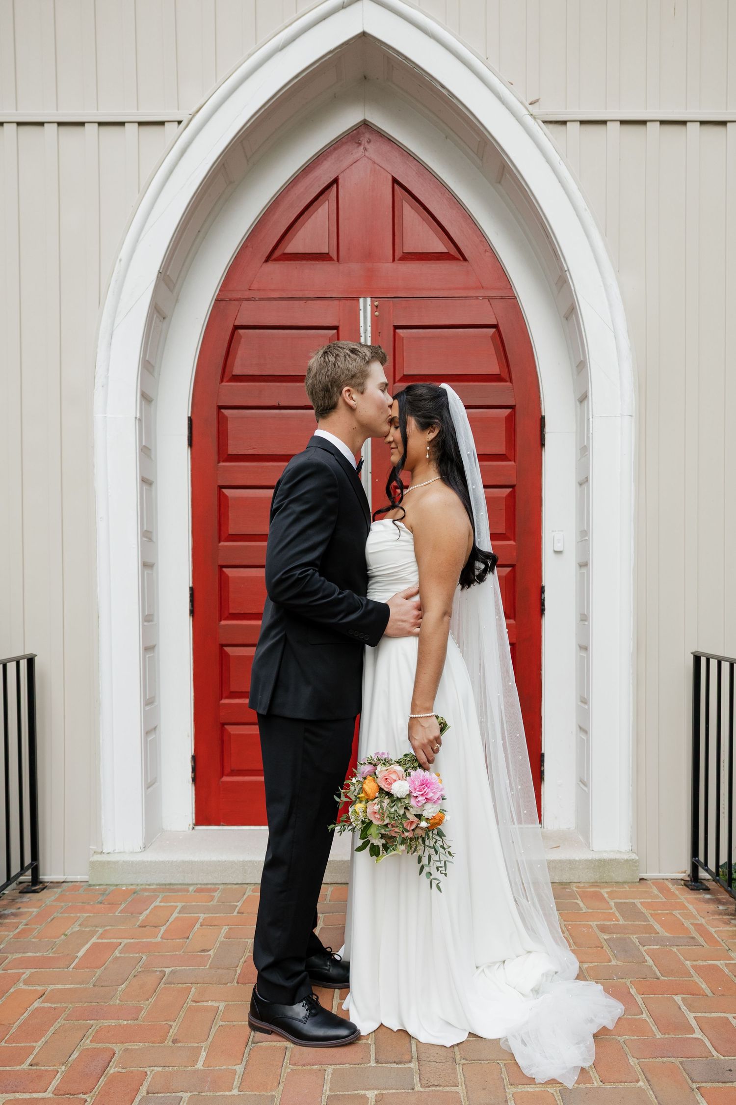 Bride and groom share a romantic almost-kiss moment in front of a stunning red arched church doorway.