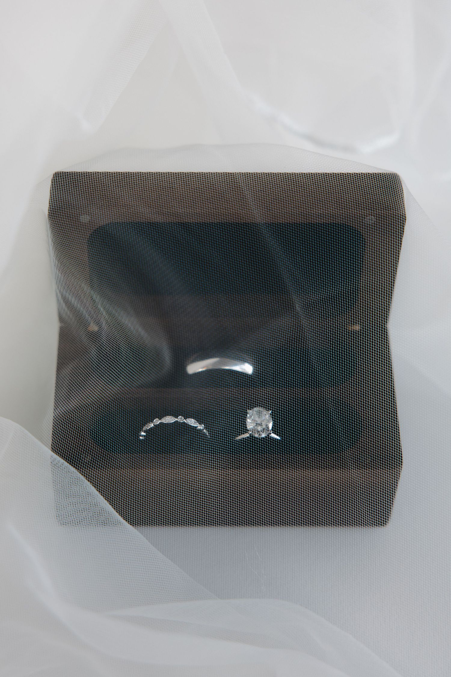 Diamond engagement ring and wedding bands displayed in a dark wooden ring box on white bridal veil.