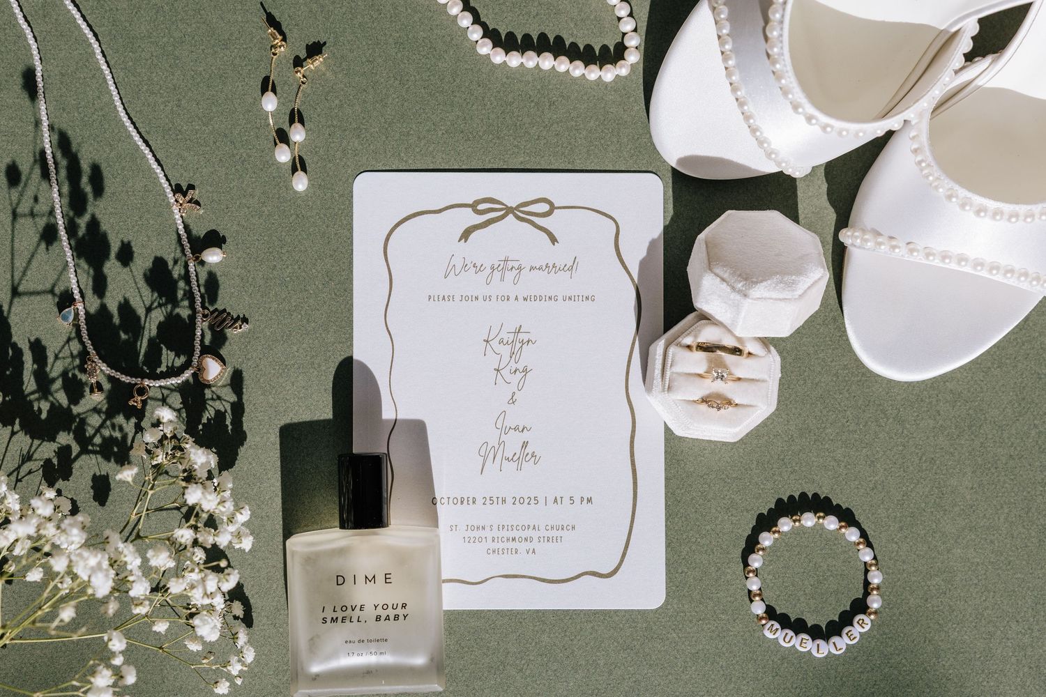 Wedding flat lay with invitation, DIME perfume, pearl jewelry, white heels, and ring box.