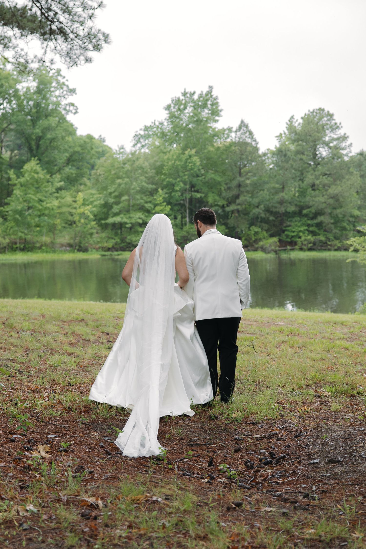 Bride and groom walking hand in hand toward a serene lake surrounded by lush green trees on their wedding day.