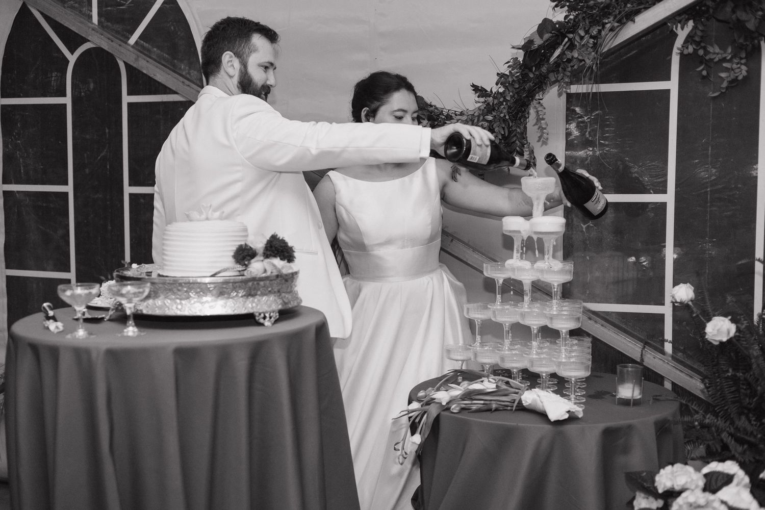 Black and white photo of bride and groom pouring champagne tower at wedding reception.