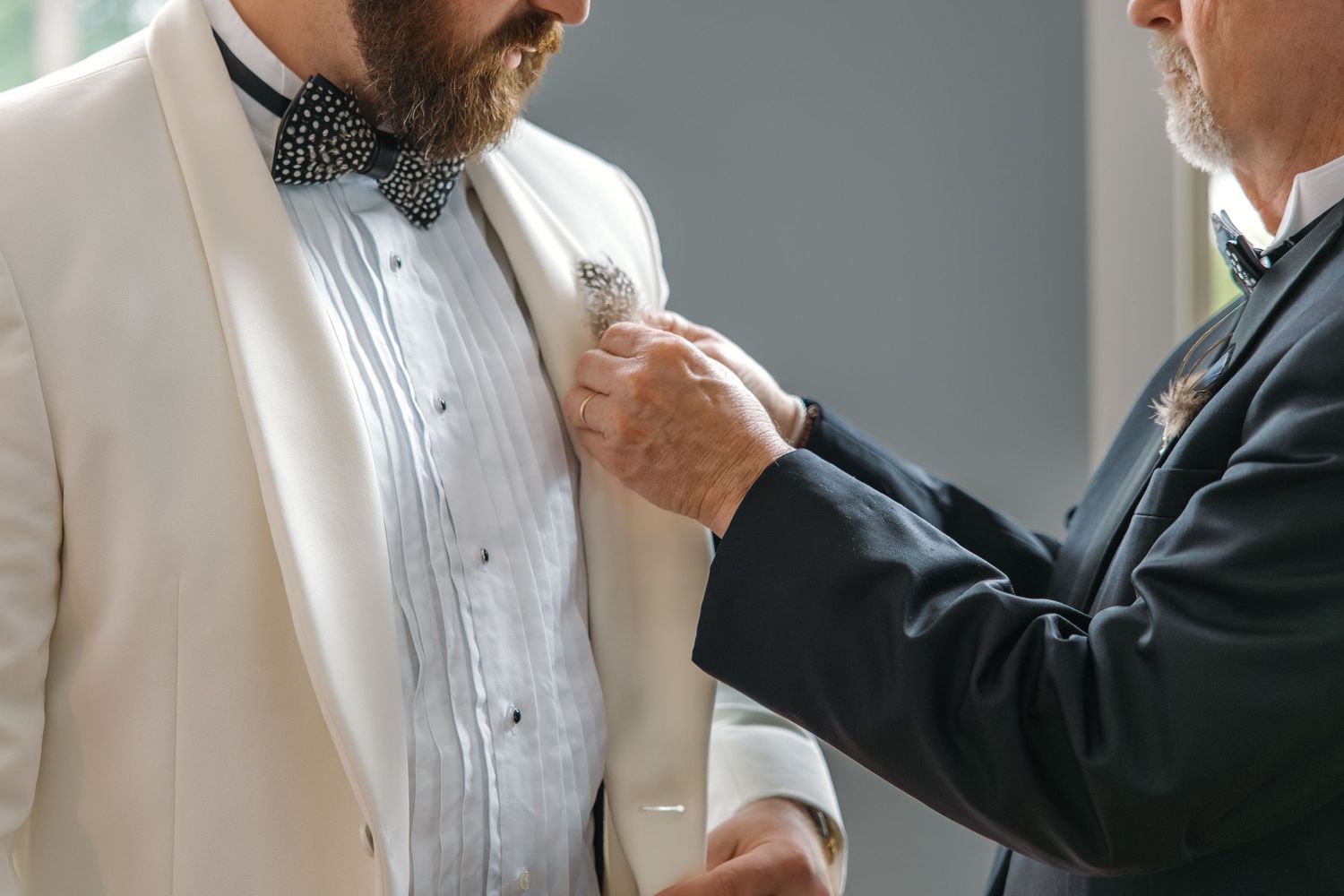 Father helping bearded groom in white tuxedo jacket adjust boutonniere before wedding ceremony.