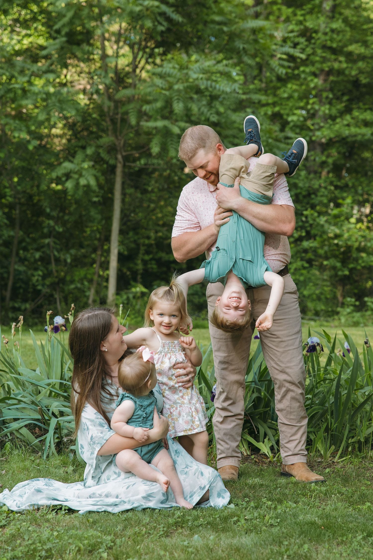 A joyful family of five plays outdoors in a lush green garden, with dad holding a laughing child upside down.