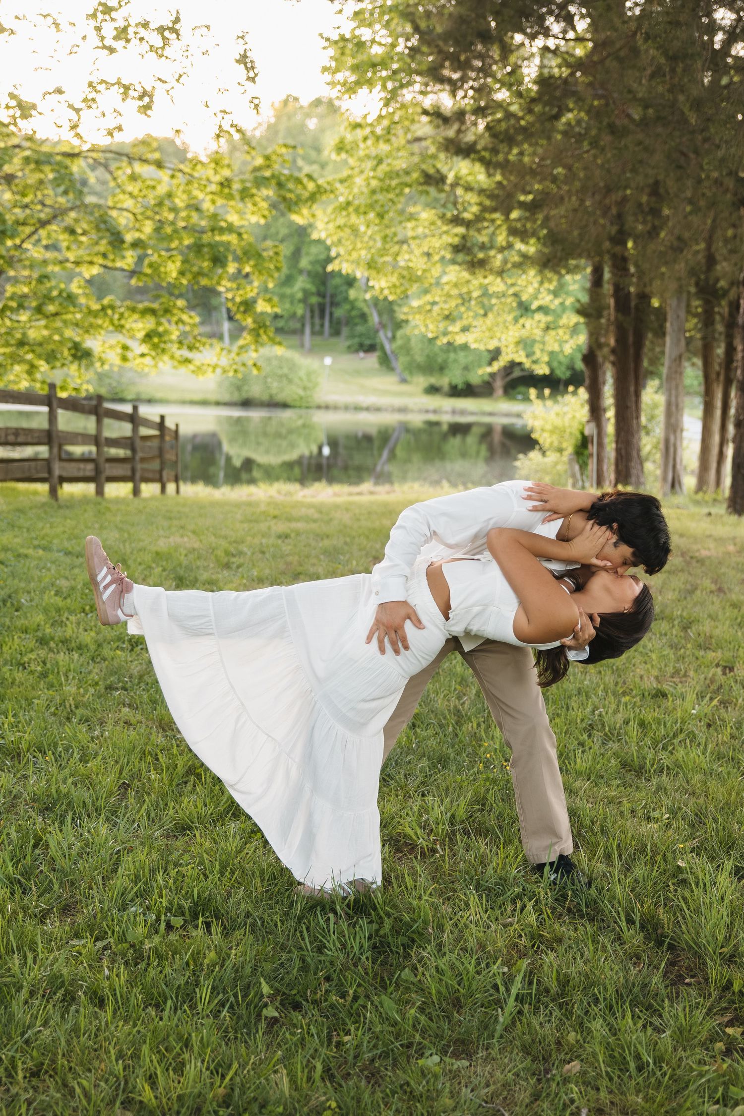 A groom dips his bride in a romantic pose on green grass with trees and a lake in the background.