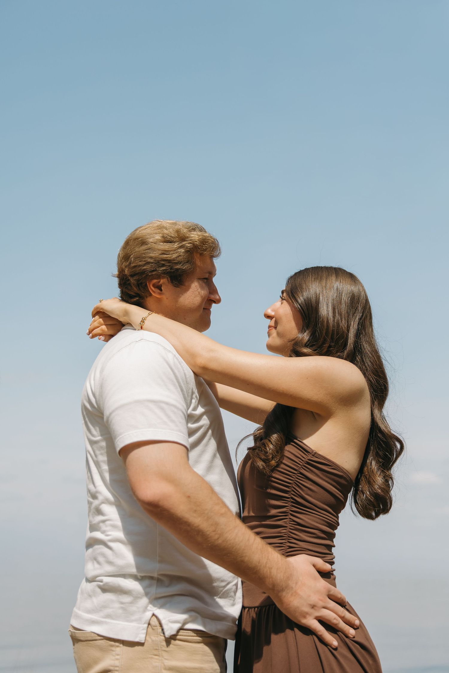 Smiling couple embracing closely against a bright blue sky in an outdoor setting.