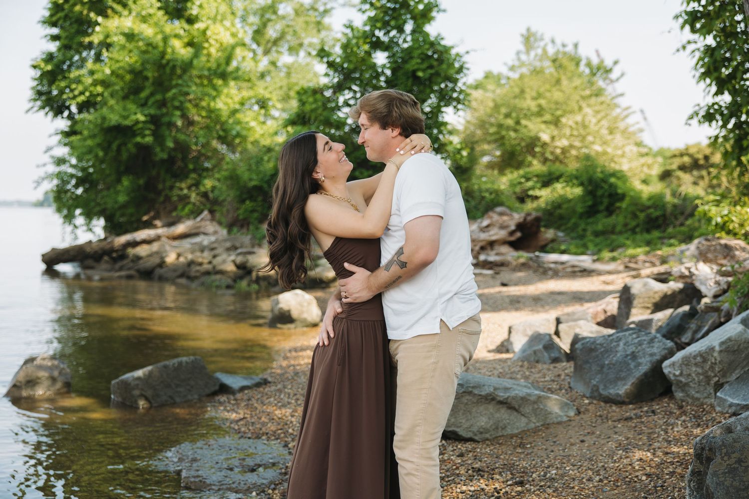 A couple shares a romantic kiss while standing on a rocky shoreline beside a calm body of water.